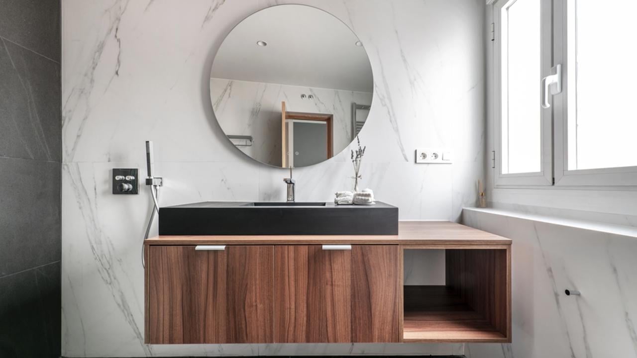 Modern design bathroom with wooden cabinets, black porcelain sink, circular mirror frame, chrome metal fixtures and fittings and black marble flooring