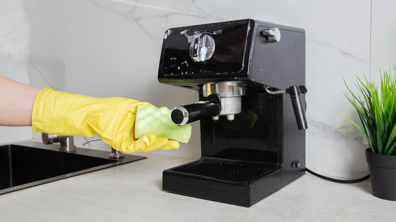 A girl s hand in a yellow glove washes a black coffee machine with a sponge with foam and cleaning agent in the kitchen against a background of a white wall with porcelain tiles. Copy space for text