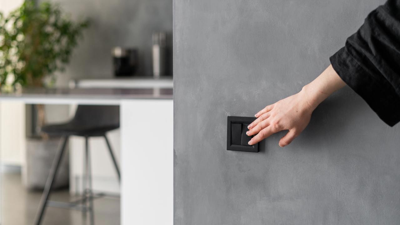 Cropped shot of woman turning light on or off in kitchen using black switch located on grey wall, modern kitchen interior design with plants, furniture and appliances in blurred background