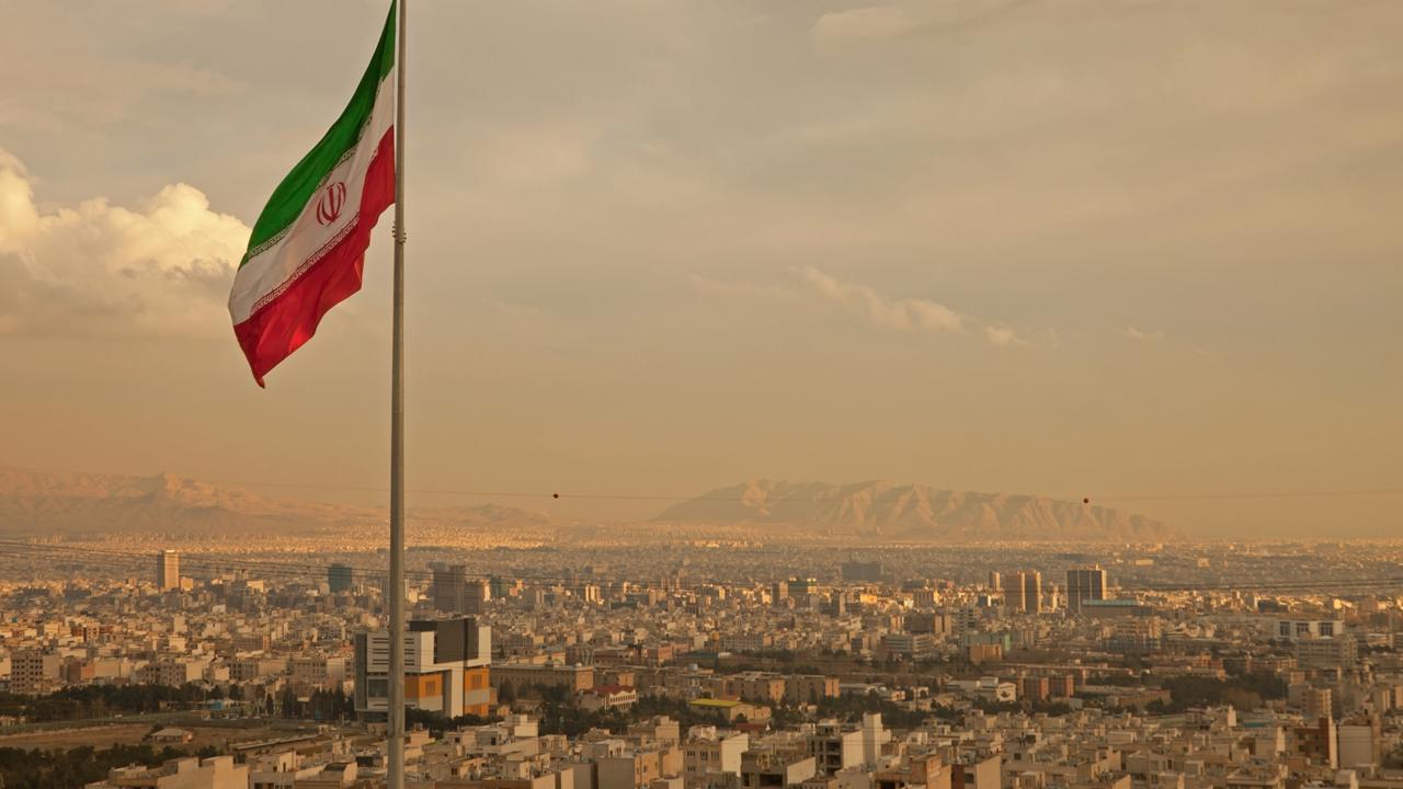 Iran flag waving  in the wind above skyline of Tehran lit by orange glow of sunset.