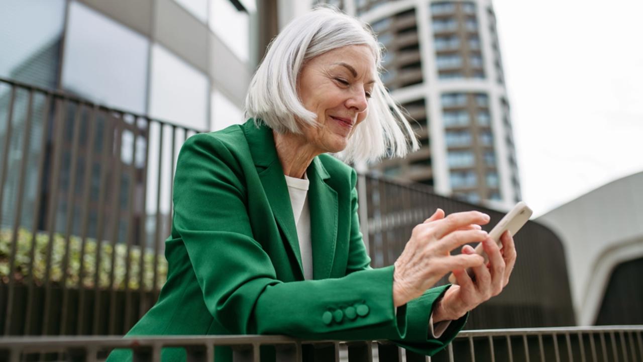 Mature businesswoman scrolling on smartphone, going home from work. Beautiful older woman with gray hair standing on city street.