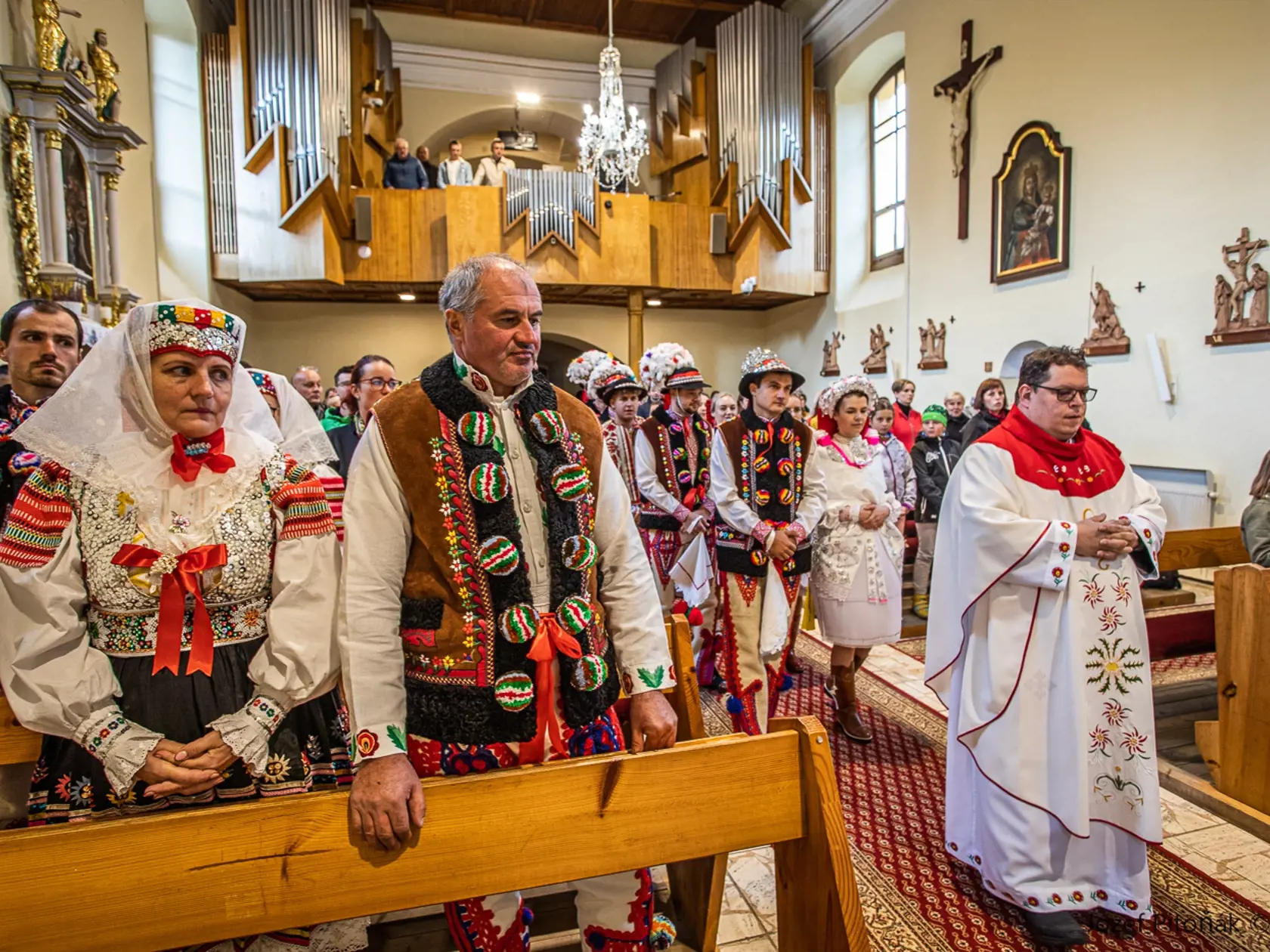 Snúbenci prichádzajú pred oltár za kňazom, ktorý následne odslúžil sviatočnú svätú omšu. Vľavo v lavici rodičia ženícha, rovnako oblečení v kroji.