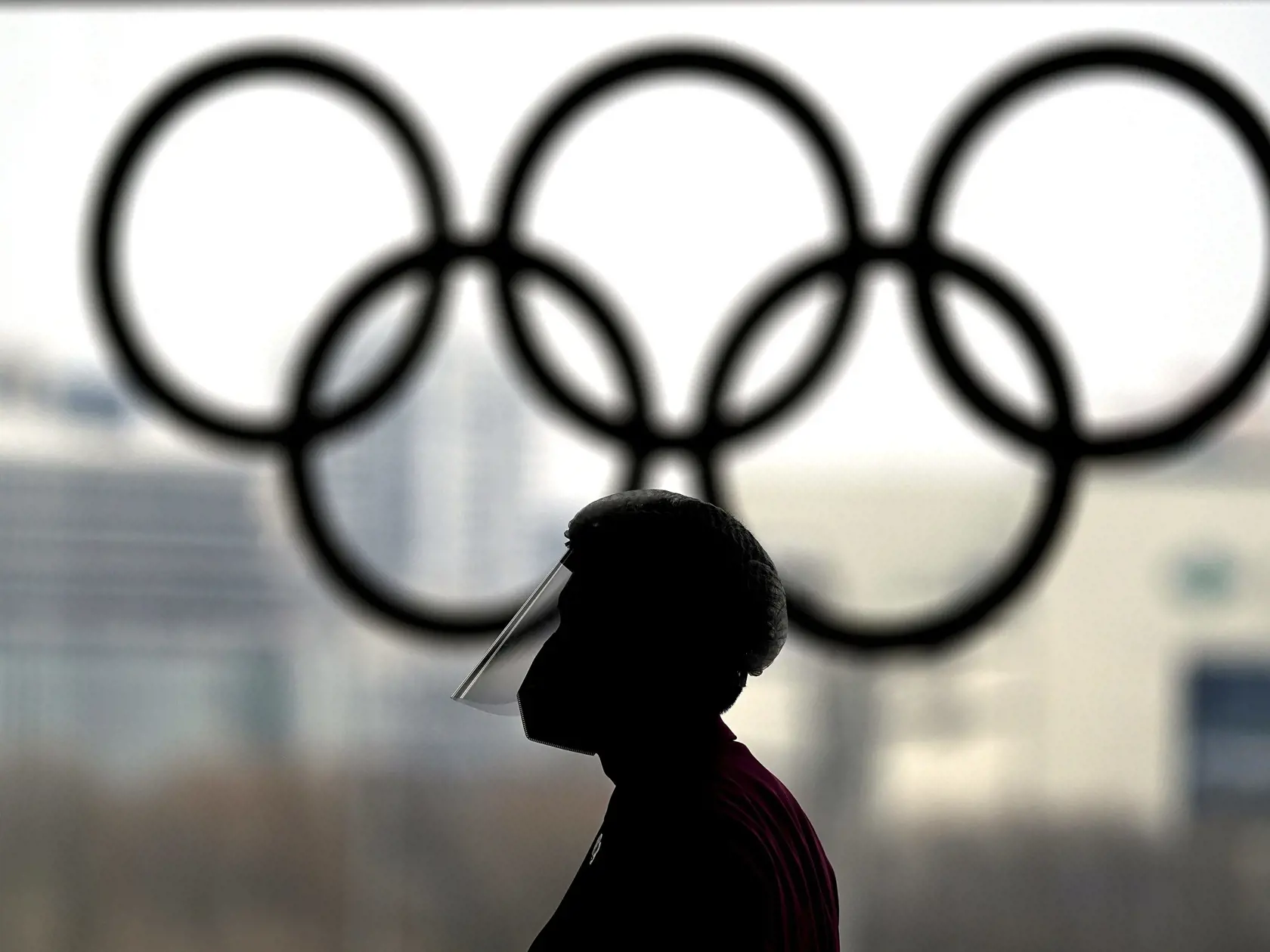 Osoba s ochranným štítom na tvári kráča okolo olympijských kruhov v novinárskom tlačovom centre na zimné olympijské hry 2022 v Pekingu 19. januára 2022. V poradí XXIV. zimné olympijské hry sa v Pekingu uskutočnia od 4. do 20. februára 2022. Paralympijské hry prídu na rad  od 4. do 13. marca.