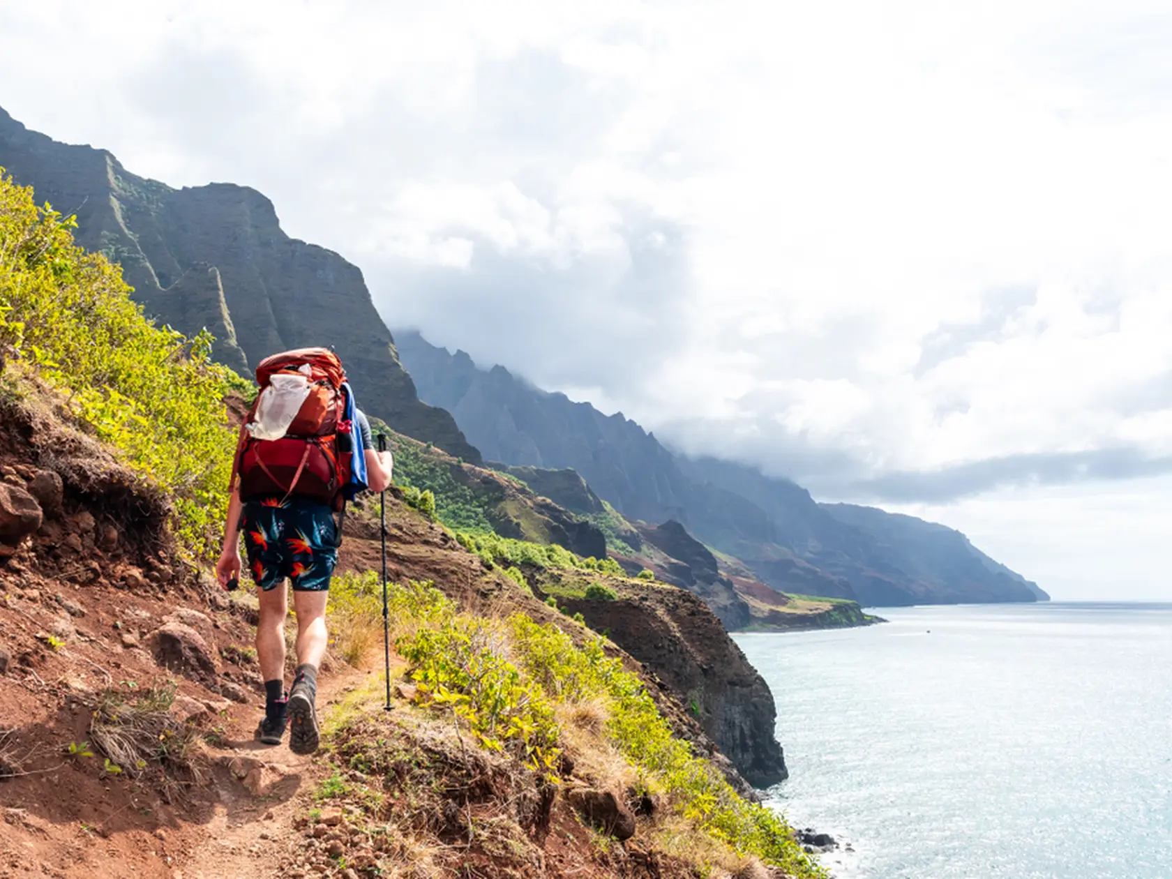 Hiker on a treacherous coastal trail with valleys and cliffs on one side and the ocean on the other. Shot on the Kalalau Trail in the Na Pali Coast State Park on the island of Kauai, Hawaii USA.