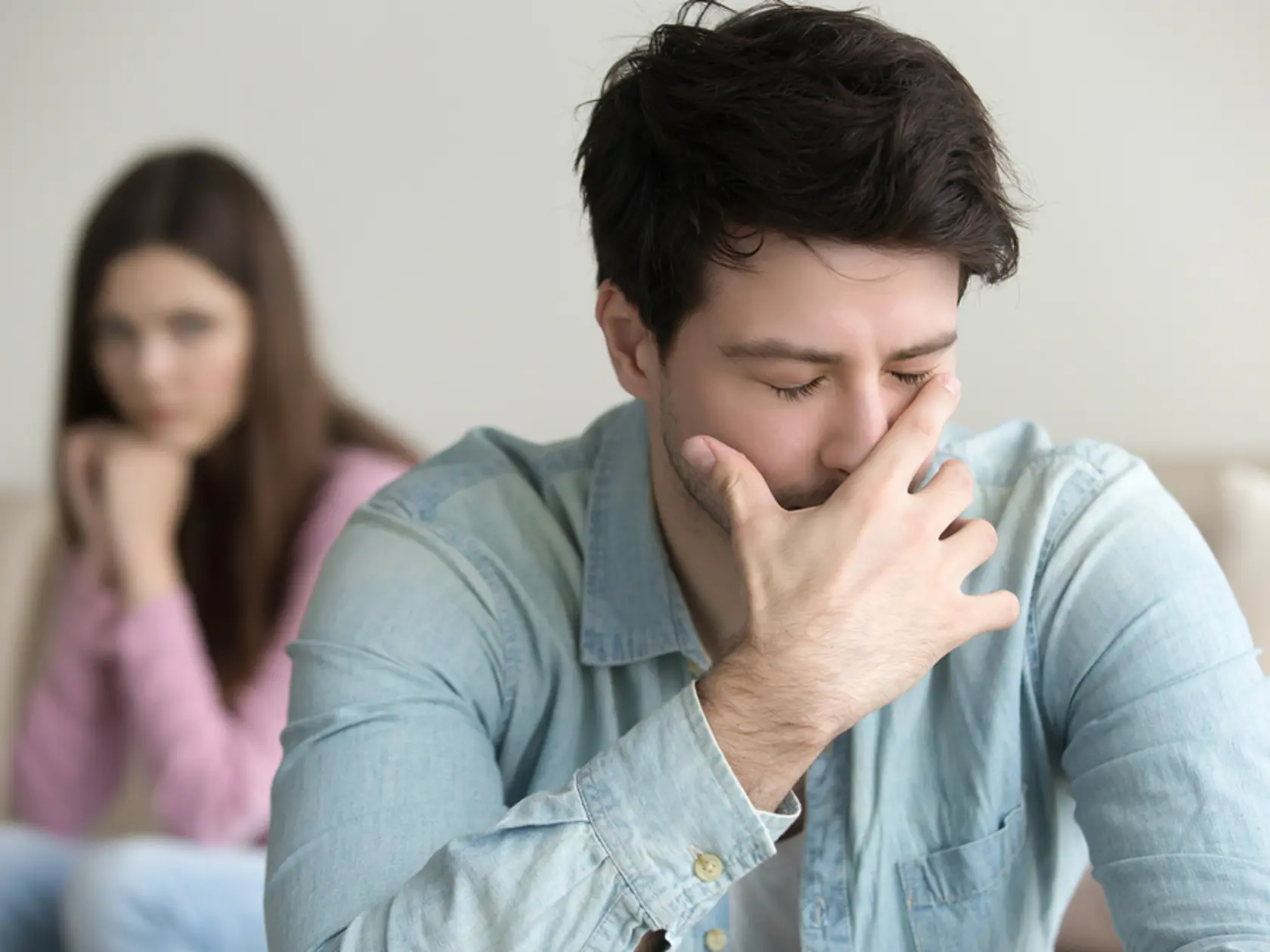 Young guy is very upset and crying, feeling guilty, depressed man in pain, girlfriend sitting in the background indoors, family relationships problems concept, businessman having troubles at work 