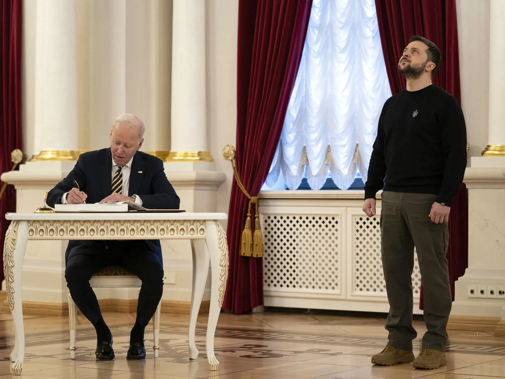 Na archívnej snímke z 20. februára 2023 americký prezident Joe Biden sa zapisuje do knihy návštev, vpravo sa prizerá ukrajinský prezident Volodymyr Zelenskyj v Kyjeve. Ukrajina a USA usporiadajú 6. a 7. decembra vo Washingtone konferenciu o vojenskom priemysle. Kyjev zvýšil úsilie o výrobu vlastných zbraní pre obavy zo zníženia dodávok zo Západu. Dúfa tiež, že spoločné podniky s medzinárodnými výrobcami zbraní pomôžu oživiť domáci priemysel.