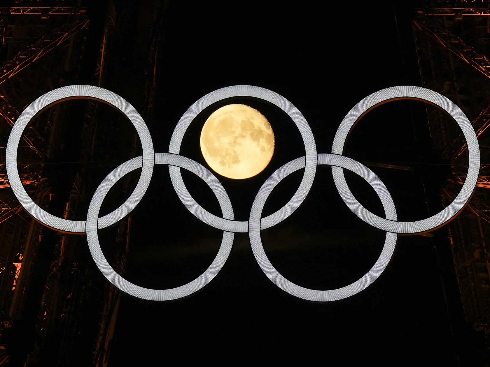 Na snímke spln mesiaca za olympijskými kruhmi, ktoré visia na Eiffelovej veži v pondelok 22. júla 2024 v Paríži.