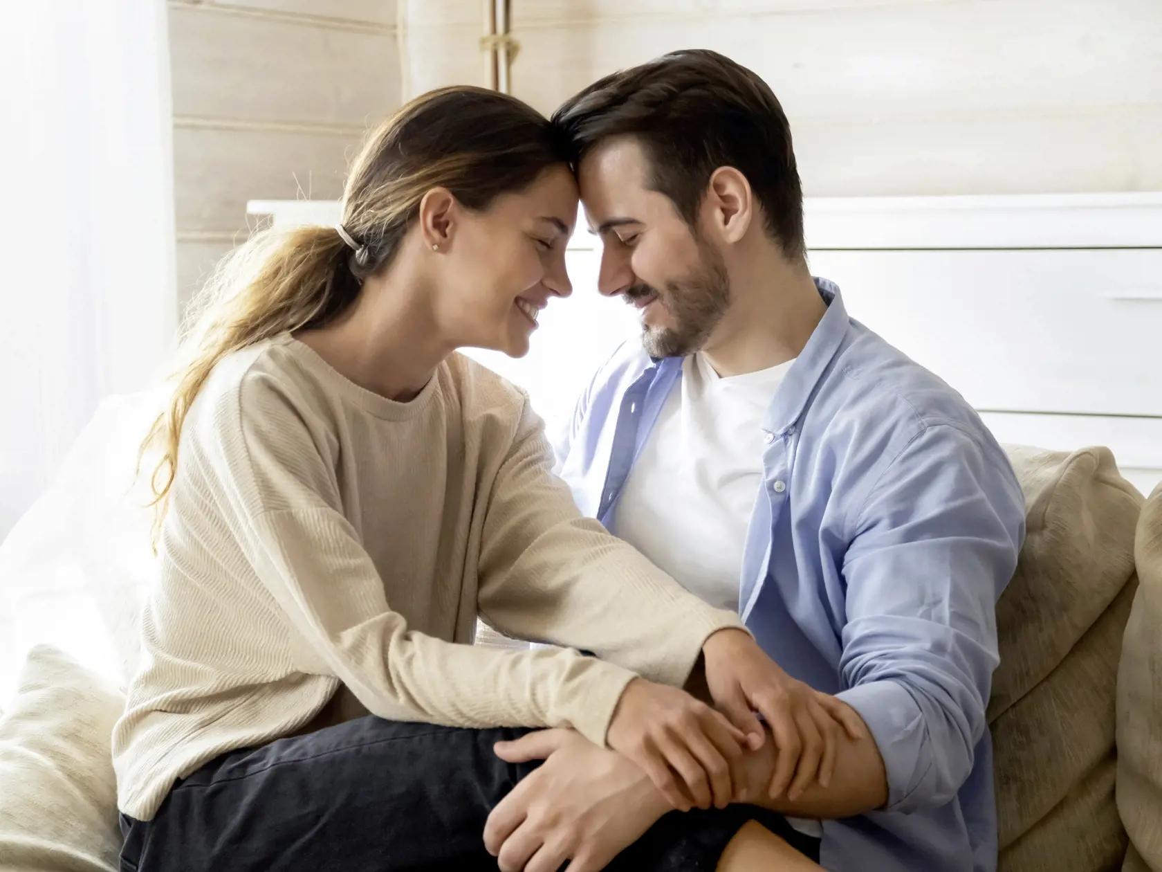 Happy loving man and woman enjoying tender moment with closed eyes, touching foreheads, expressing unity and care, beautiful girlfriend sitting on boyfriend laps, relaxing on cozy couch at home