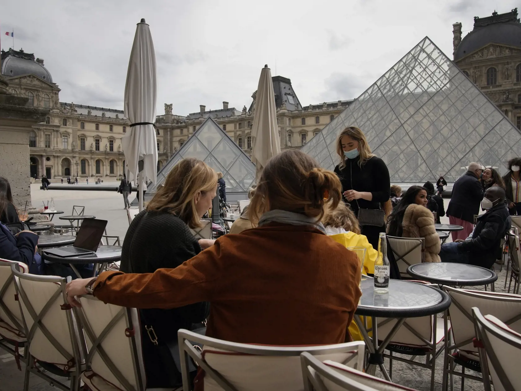 Ľudia sedia na terase kaviarne pred Múzeom Louvre v Paríži.