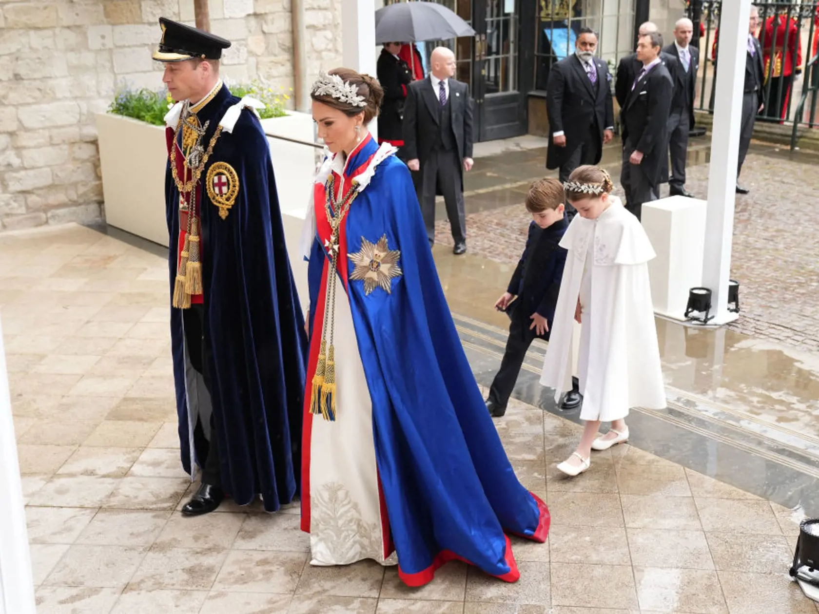 Princezná Kate mala na sebe tradičný ceremoniálny plášť. Pod ním slonovinové šaty od Alexandra McQueena a 3D čelenku, rovnako tak aj jej dcéra, princezná Charlotte.