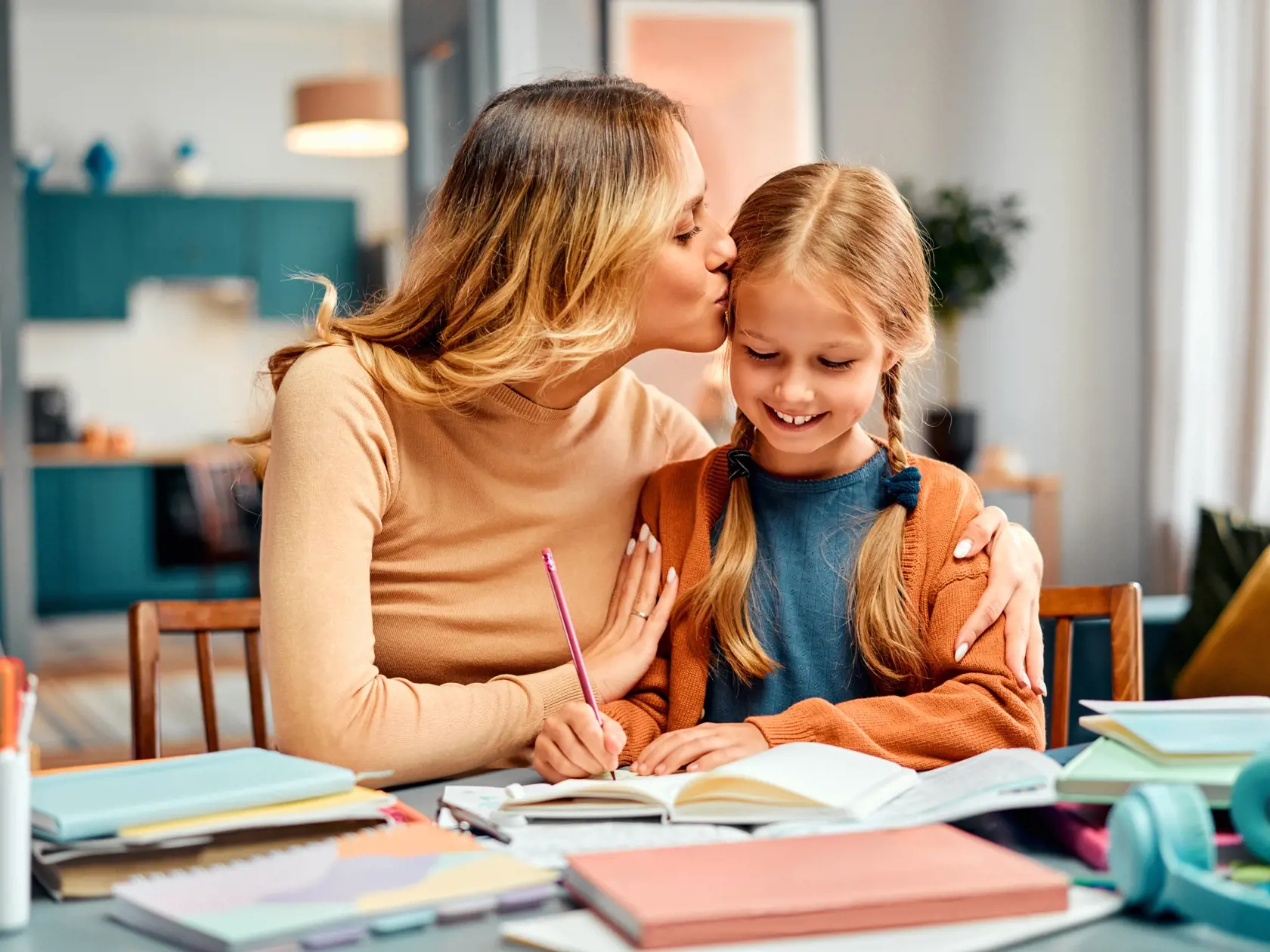 Concept of motherhood. Loving female kissing daughter in forehead in living room. Smiling girl receiving dose of motivation from mum and keep trying solving task in daybook.