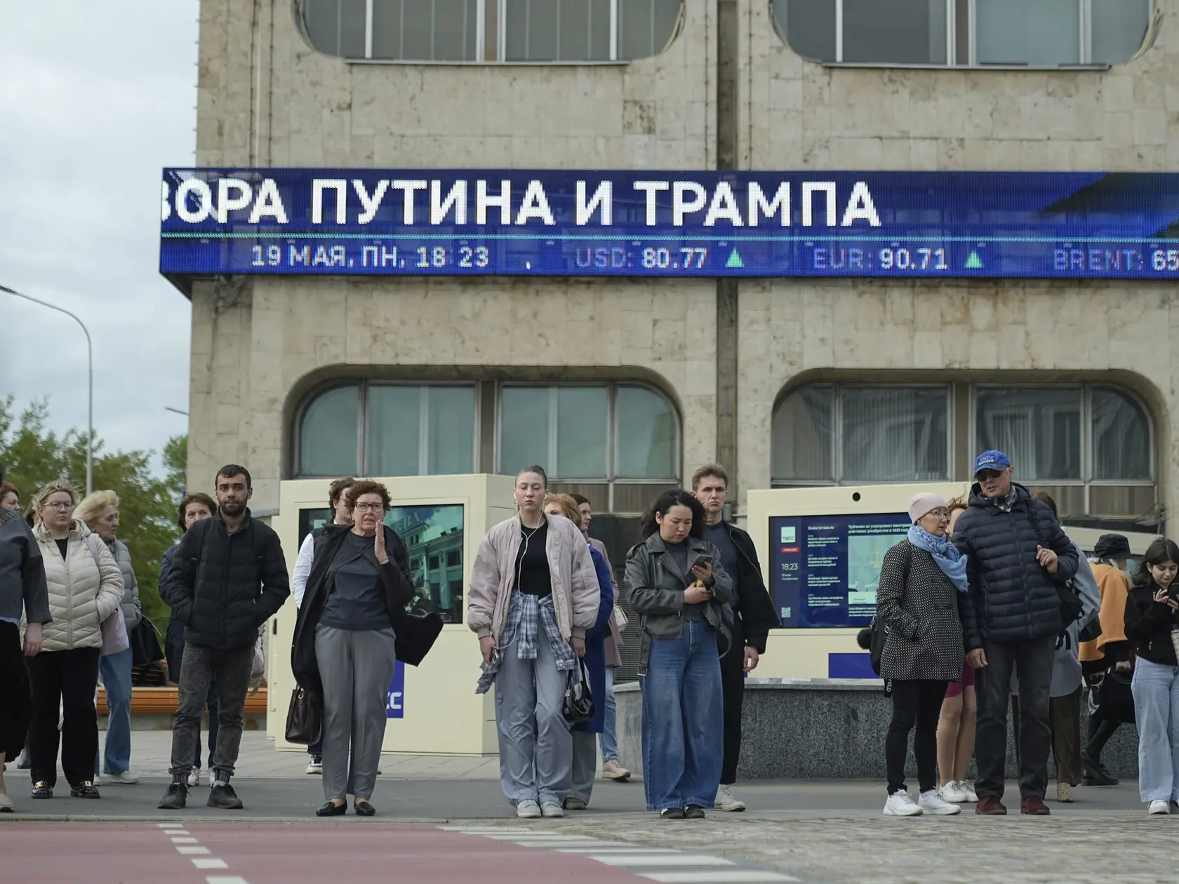 Ľudia kráčajú cez cestu v Moskve, v pozadí ticker informuje o telefonáte medzi americkým prezidentom Donaldom Trumpom a ruským prezidentom Vladimirom Putinom v pondelok 19. mája 2025.