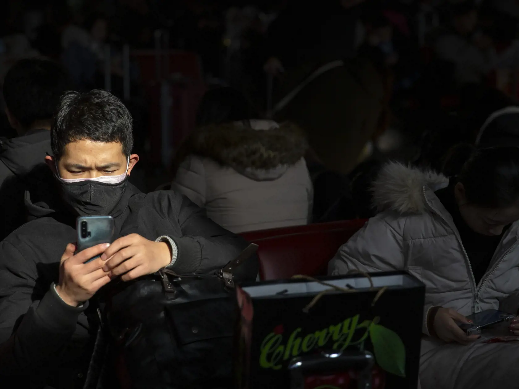 Cestujúci, ktorý má na tvári ochranné rúško, sedí v čakárni a pozerá do mobilného telefónu na železničnej stanici v Pekingu.
