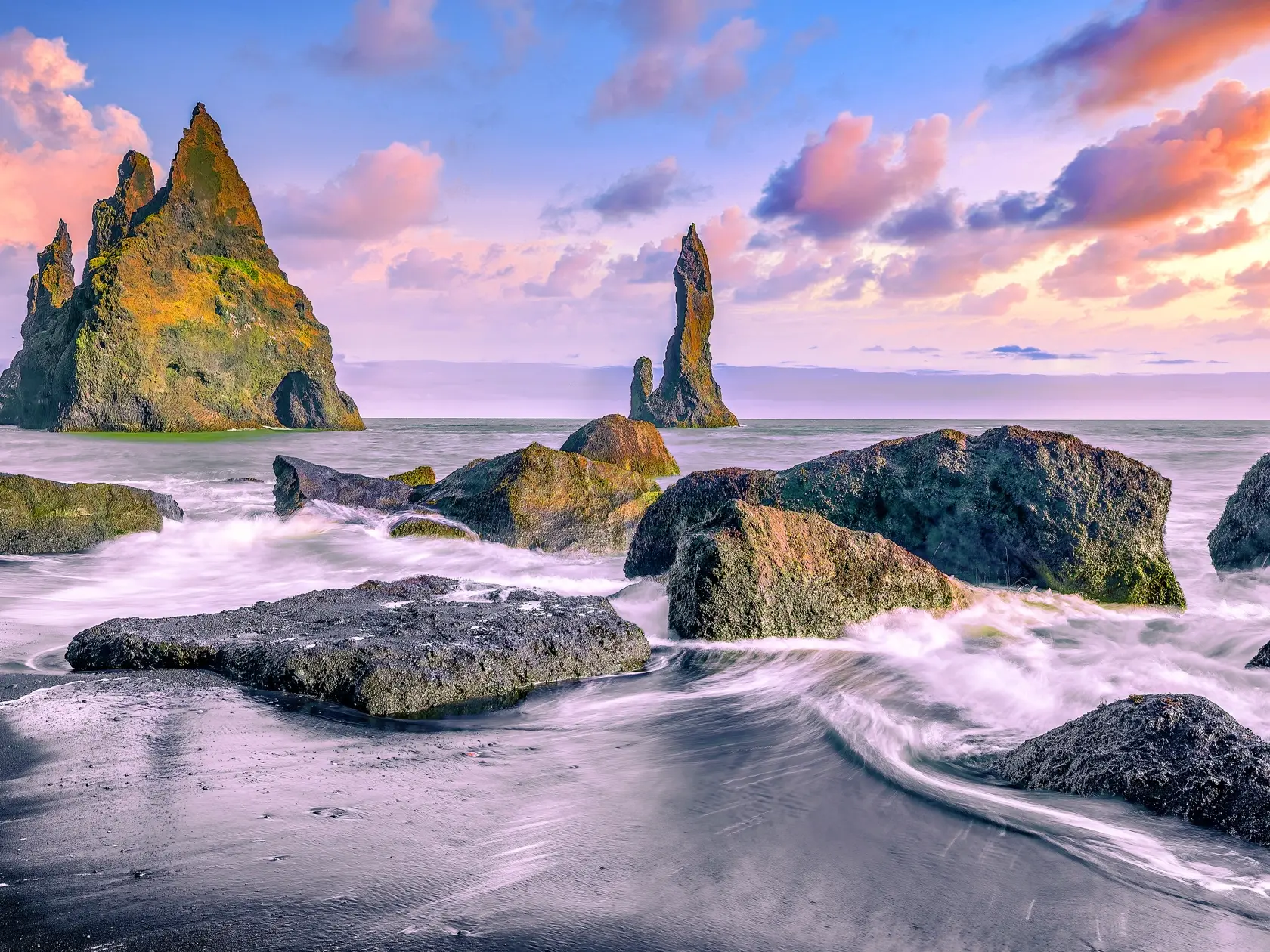 Čadičové skaly – Reynisfjara. Mohutné tmavé stĺpy čadiča sa týčia z čiernej piesočnatej pláže, kde rozbúrené vlny Atlantiku narážajú na monumentálne skalné útvary, vytvárajúc kontrast medzi silou prírody a krásou pobrežia.