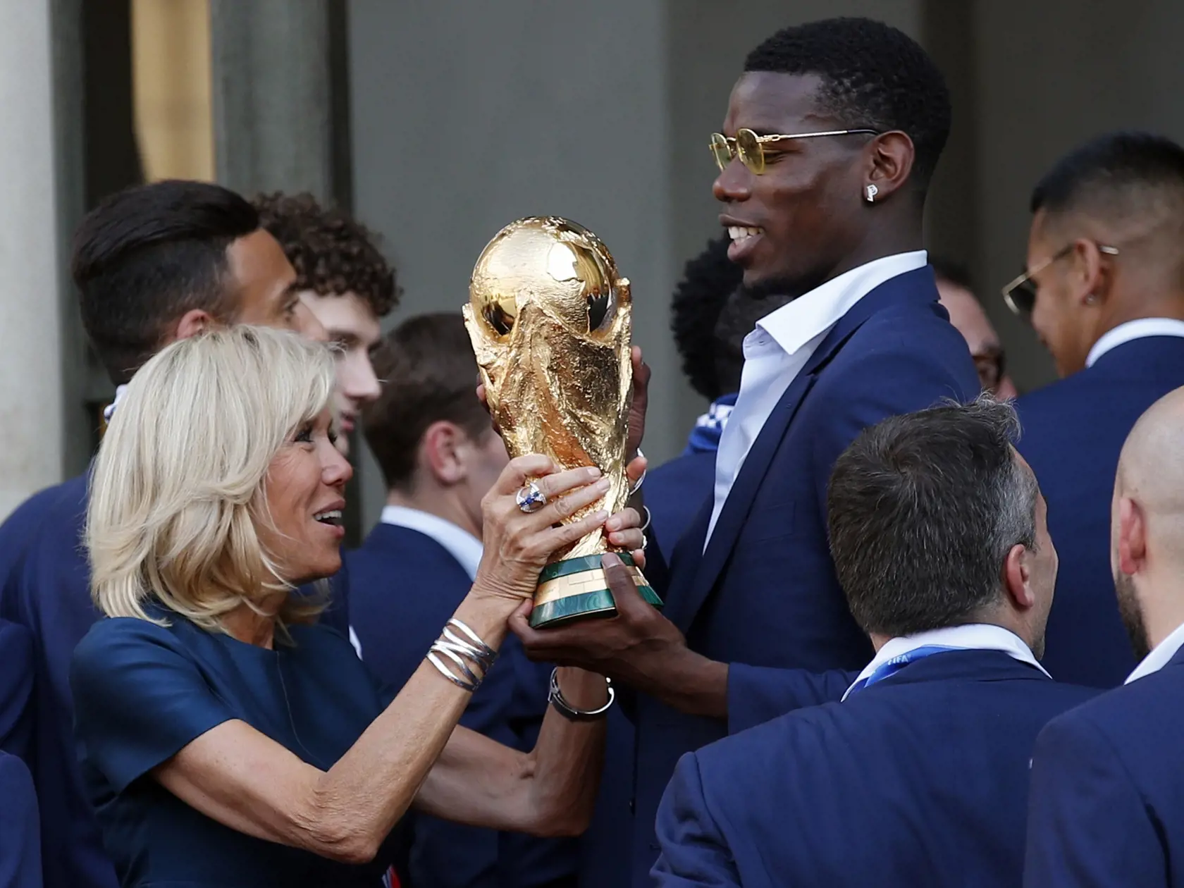 SY 72 Paríž - Na snímke francúzsky futbalový reprezentant Paul Pogba (vpravo) pomáha zdvihnúť trofej majstrov sveta manželke francúzskeho prezidenta Brigitte počas prijatia u francúzskeho prezidenta Emmanuela Macrona po príchode do Paríža 16. júla 2018. FOTO TASR/AP