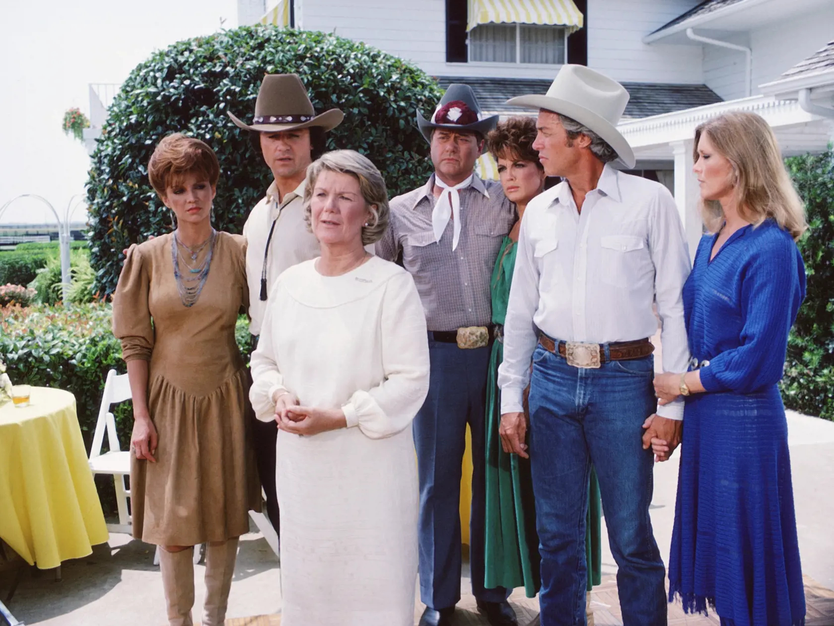 In a scene from the American television series 'Dallas, cast members gather on the porch of a large house, Dallas, Texas, 1981. From left, American actors Victoria Principal (as Pamela Barnes Ewing), Patrick Duffy (as Bobby Ewing), Barbara Bel Geddes (as Eleanor Southworth 'Miss Ellie' Ewing) (foreground), Larry Hagman (as John Ross 'J.R.' Ewing, Jr.), Linda Gray (as Sue Ellen Ewing), Steve Kanaly (as Ray Krebbs), and Susan Howard (as Donna Culver). (Photo by CBS Photo Archive/Getty Images)