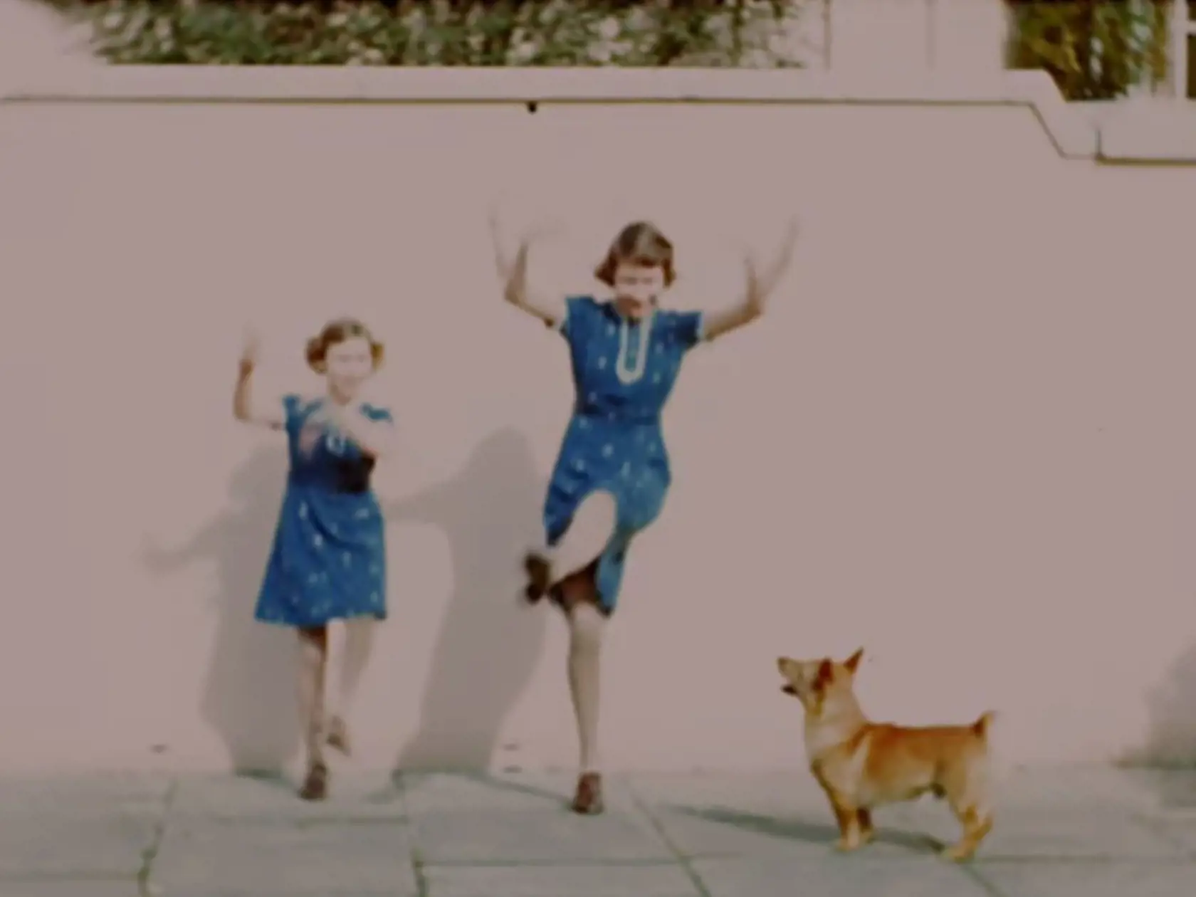 Princezná Margaret a princezná Elizabeth tancujú v Royal Lodge Windsor v roku 1940 a vedľa nich je corgi.