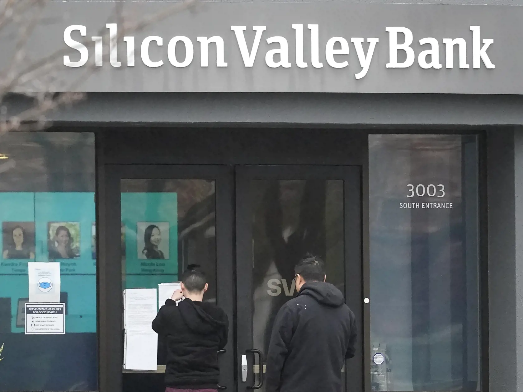 KK35 Santa Clara - Ľudia stoja pred vstupom do banky Silicon Valley Bank 10. marca 2023 v Santa Clare.  Americké úrady v piatok zatvorili Silicon Valley Bank (SVB), aby ochránili vkladateľov. V pondelok (13. 3.) sa jej pobočky znovu otvoria, ale pod kontrolou federálneho úradu na ochranu vkladov (FDIC). FOTO TASR/AP
People look at signs posted outside of an entrance to Silicon Valley Bank in Santa Clara, Calif., Friday, March 10, 2023. The Federal Deposit Insurance Corporation is seizing the ass