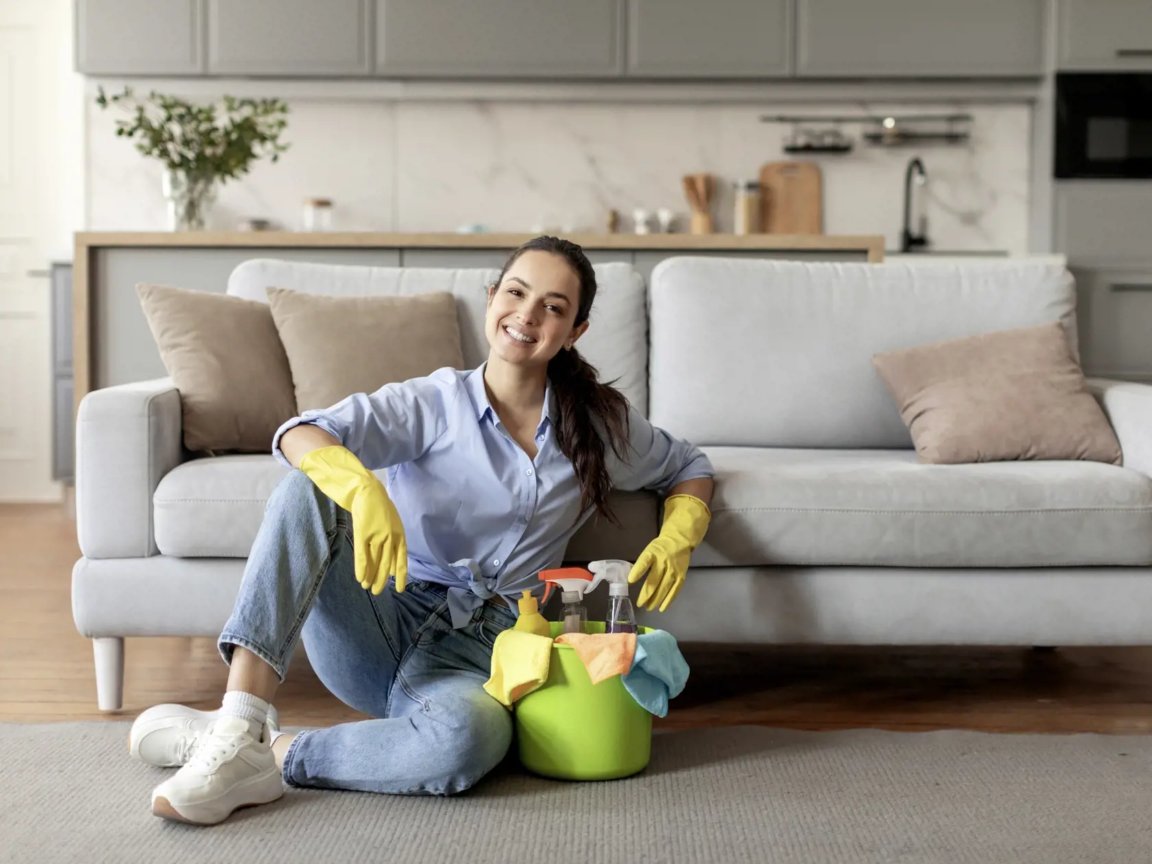 Content woman in casual attire and yellow gloves sitting on the floor, taking break with bucket of cleaning supplies next to her, living room interior, free space