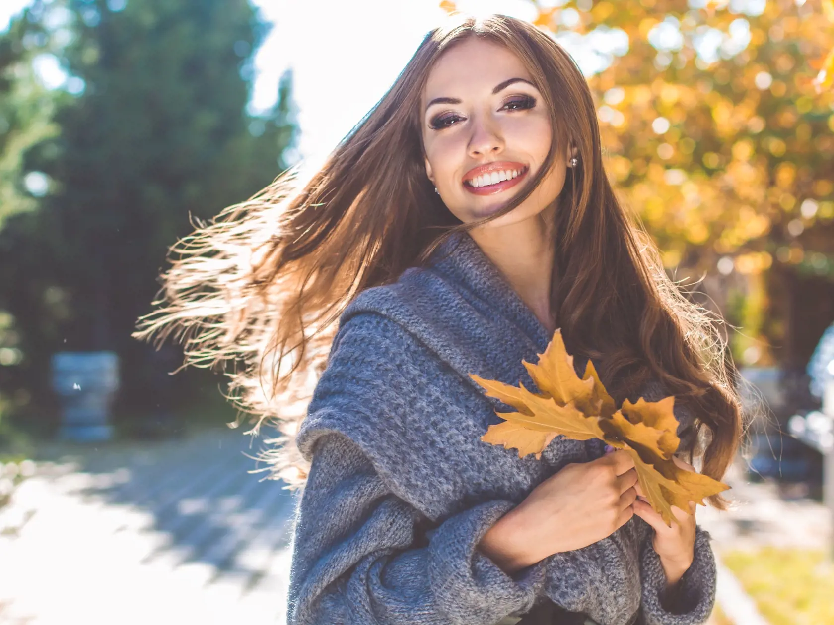 Happy girl is wearing warm knitted gray coat in park with maple yellow leaves, autumn time