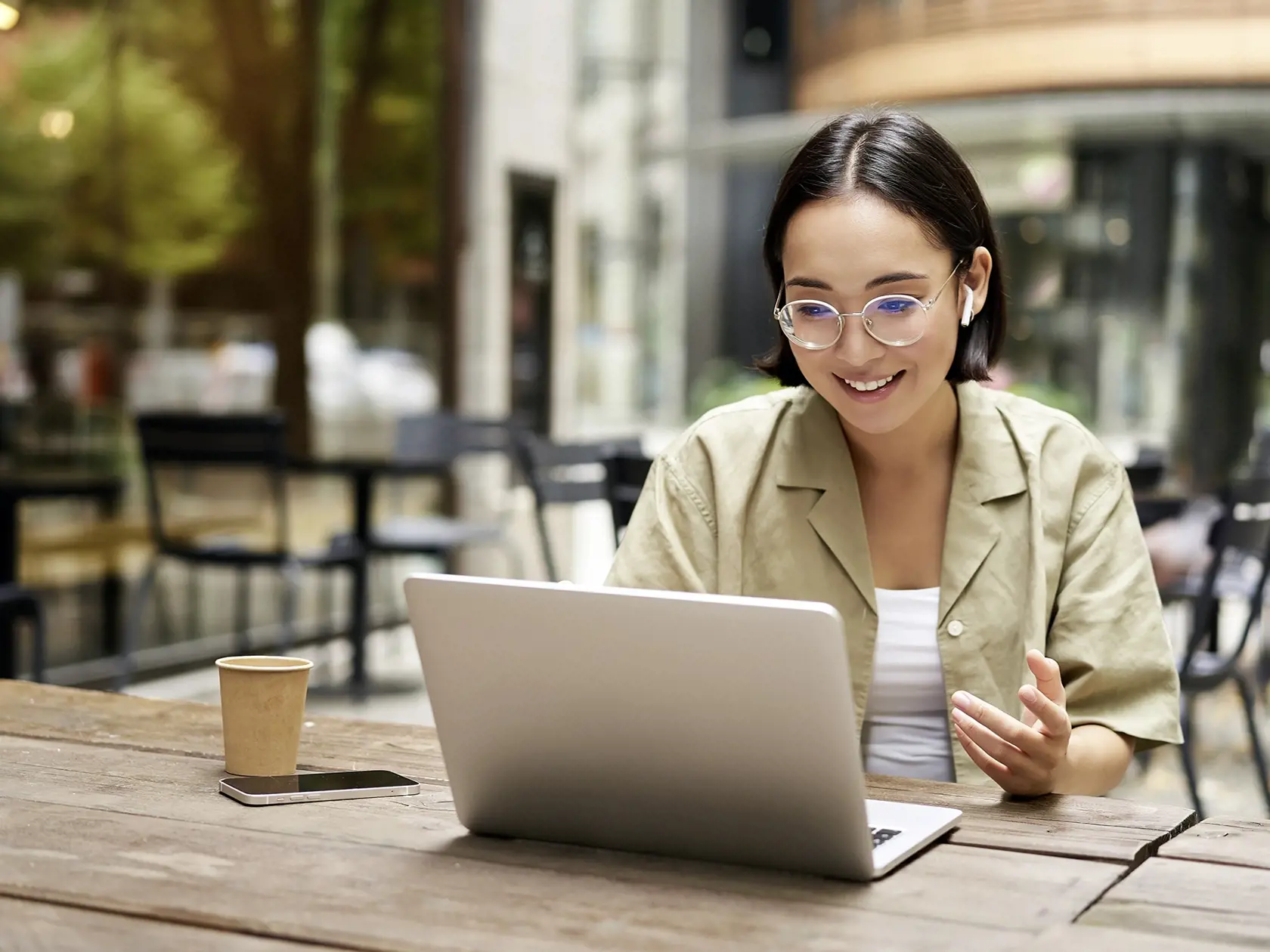 Young woman sitting on online meeting in outdoor cafe, talking to laptop camera, explaining something, drinking coffee.