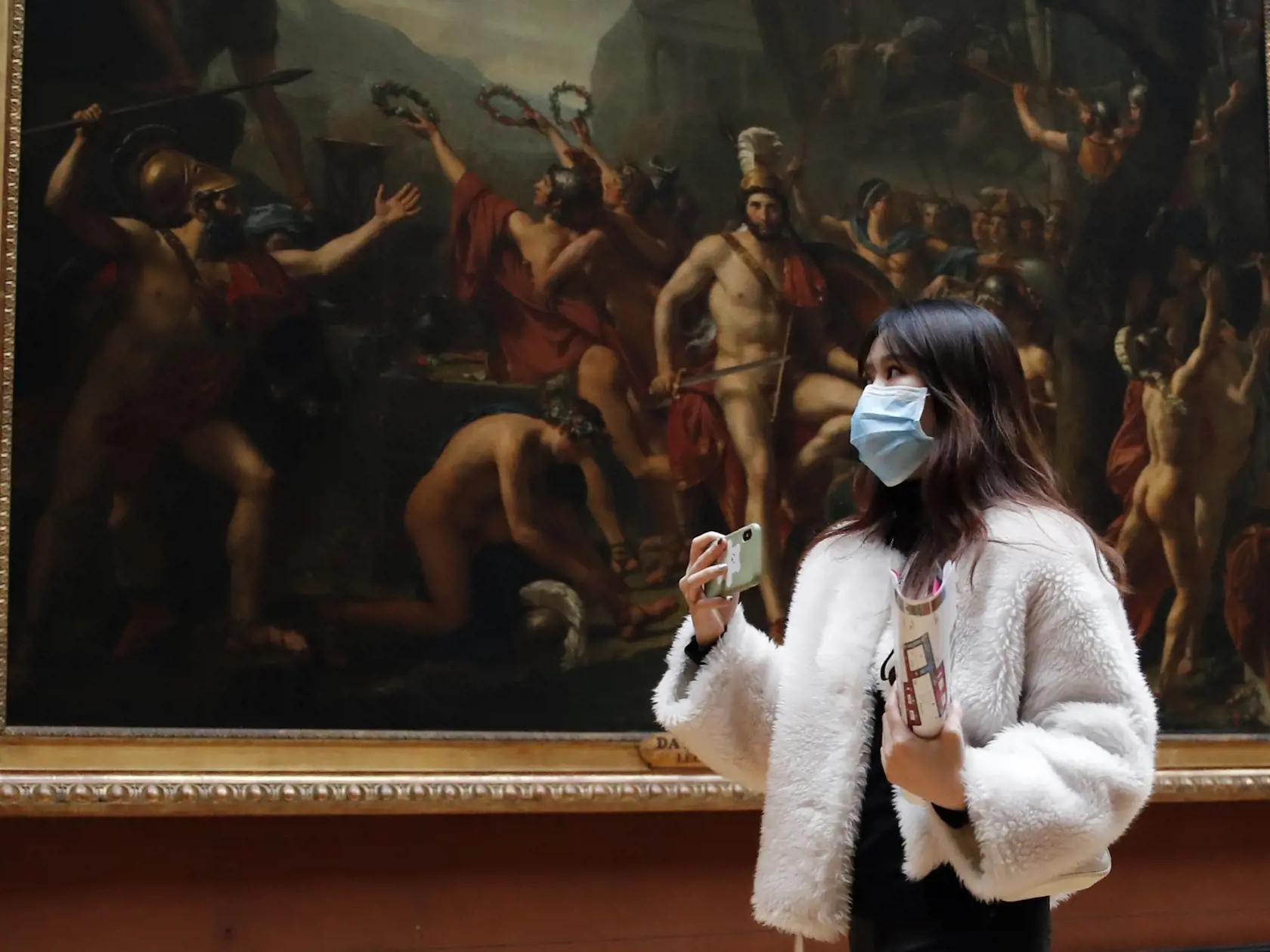 Žena s ochranným rúškom stojí pred olejom na plátne z roku 1814 s názvom Leonidas v Termopylách od Jacques-Louis Davida v múzeu Louvre v Paríži 5. marca 2020.