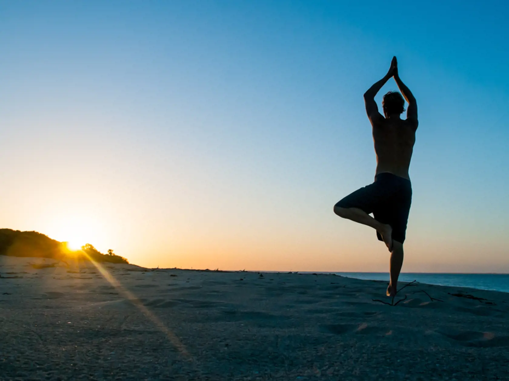 Fit young man practices sun salutation yoga on the beach at sunset