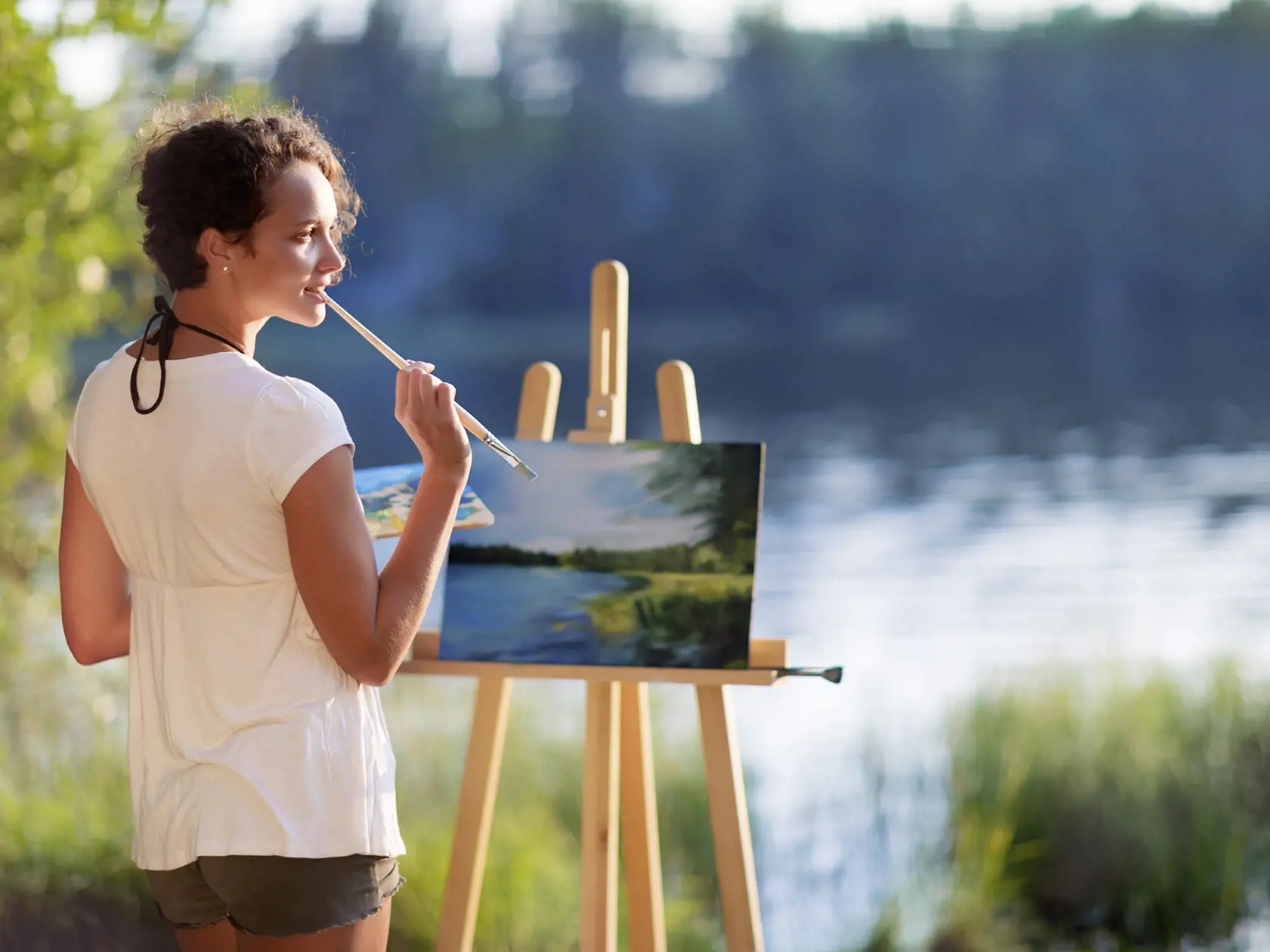 Young pretty woman artist draws paints a picture of a lake on open plain air outdoors