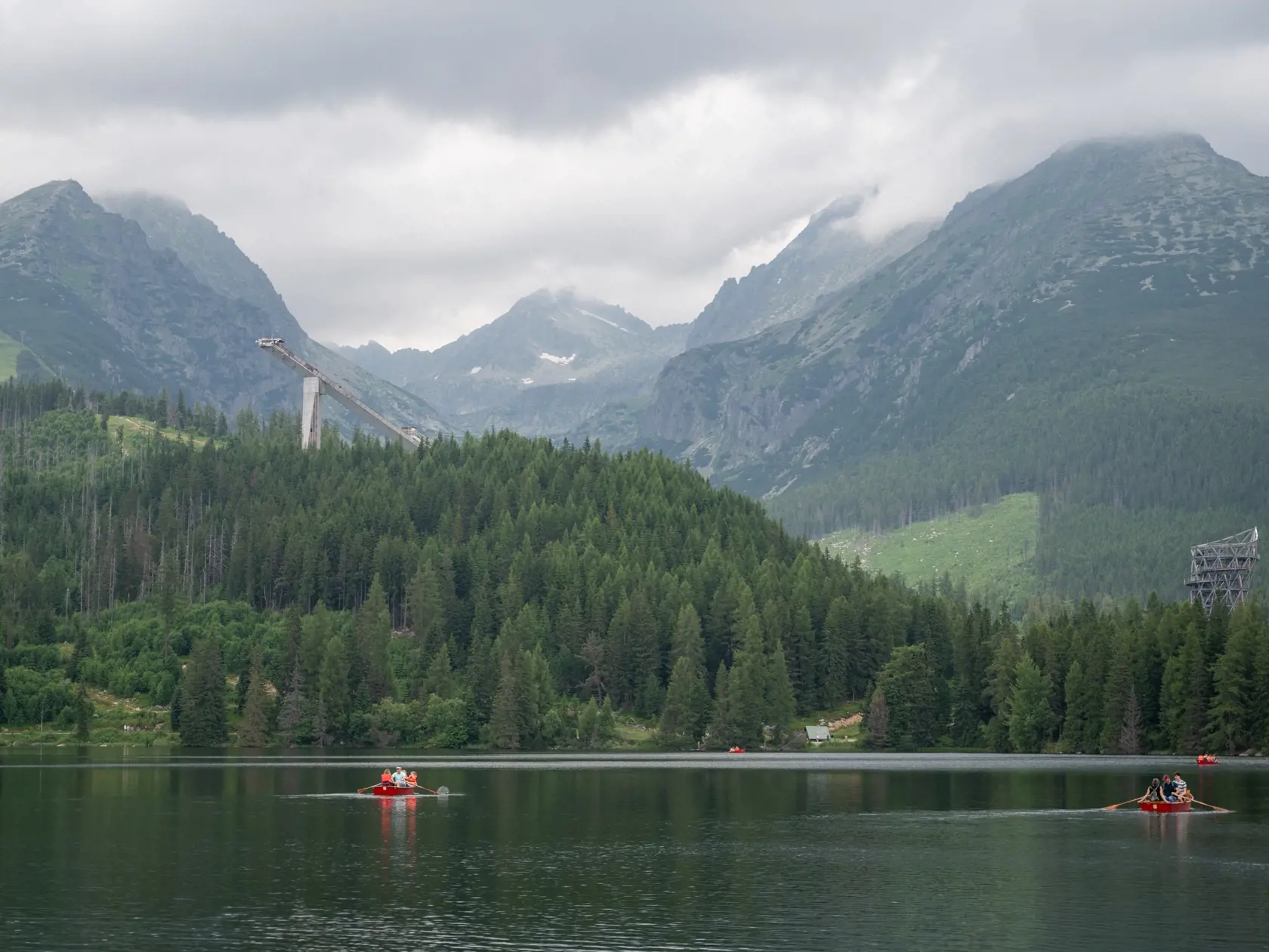 Turisti sa člnkujú na Štrbskom plese, v pozadí zľava vrchy Predné Solisko, Hlinská veža a Patria,  v popredí vľavo  skokanský mostík a v popredí vpravo vyhliadková veža počas letnej turistickej sezóny na Štrbskom Plese.