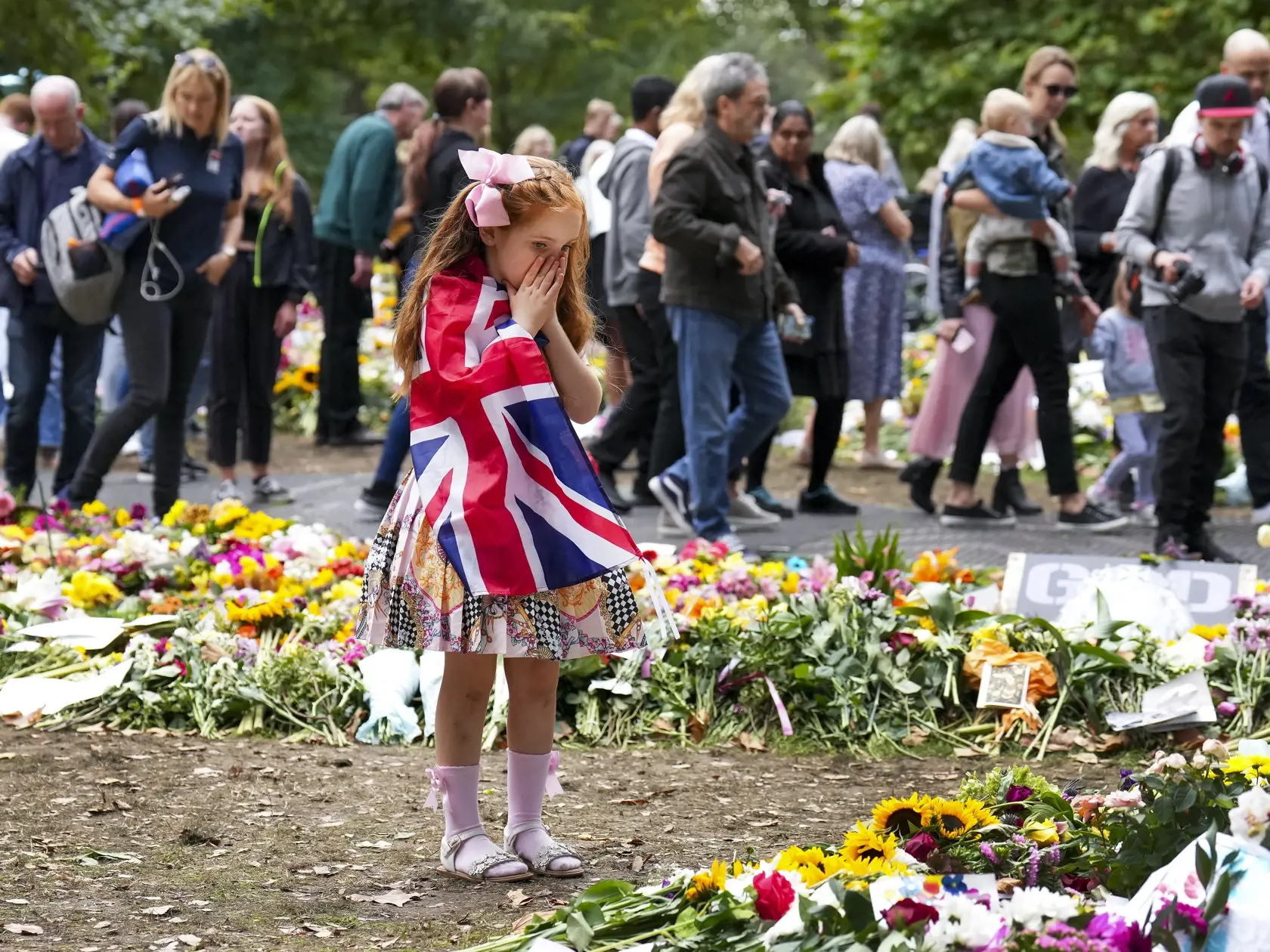Na snímke v popredí šesťročná Ann Doranová odetá do britskej zástavy si zakrýva tvár pri pohľade na množstvo kvetov, ktoré zanechali na pamiatku zosnulej britskej kráľovnej Alžbety II. smútiaci ľudia pred bránami Buckinghamského paláca v Londýne 15. septembra 2022.