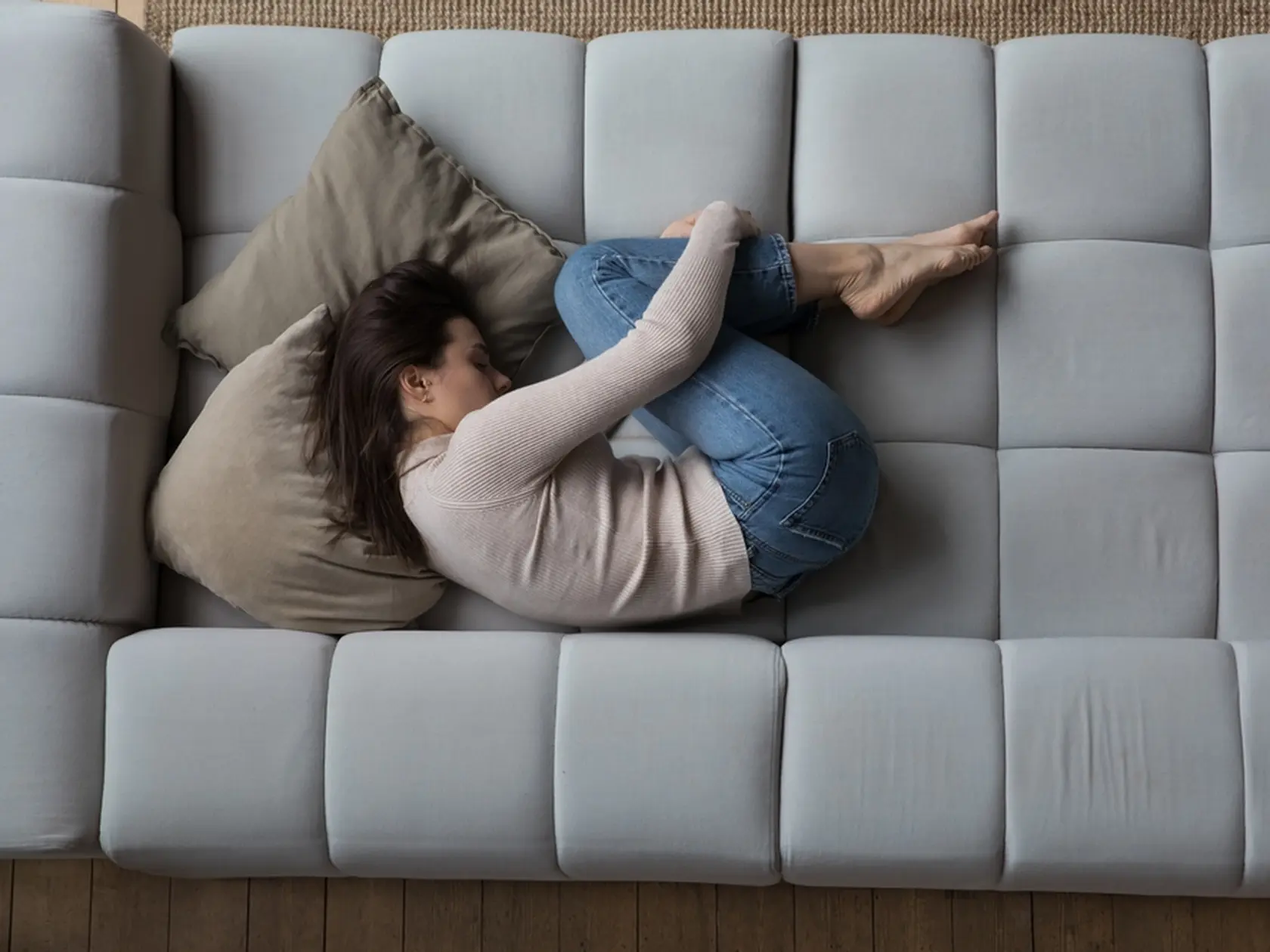 Frustrated depressed millennial girl lying in embryo pose on couch, sleeping, drowsing, feeling stress, apathy, suffering from abdominal pain, bellyache. Depression concept. Top view