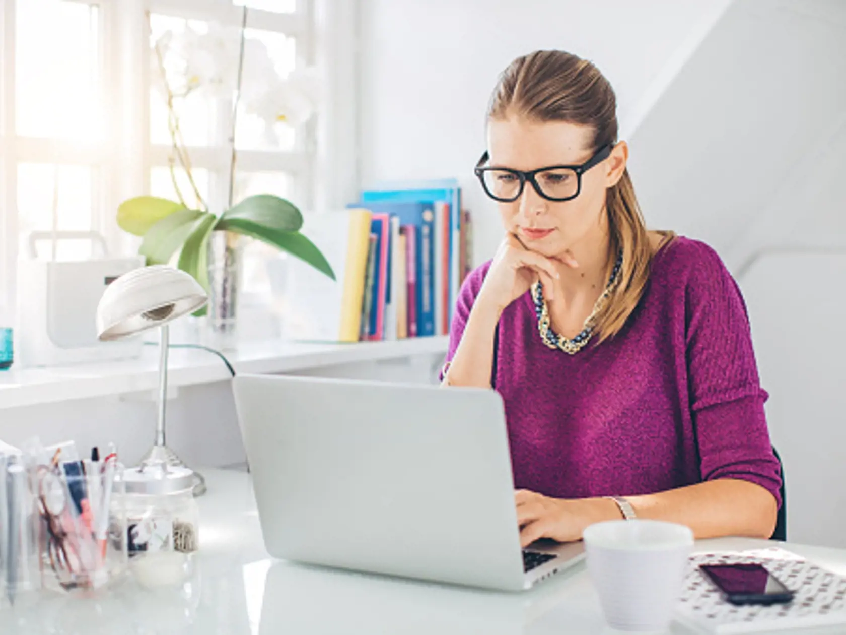 Woman working in her attic home office, with big window and natural light. Sitting at the table, wearing glasses and elegant clothes. Laptop is on the table. Hands near face. Office colour is grayish white. Nordic style. Copenhagen, Denmark.