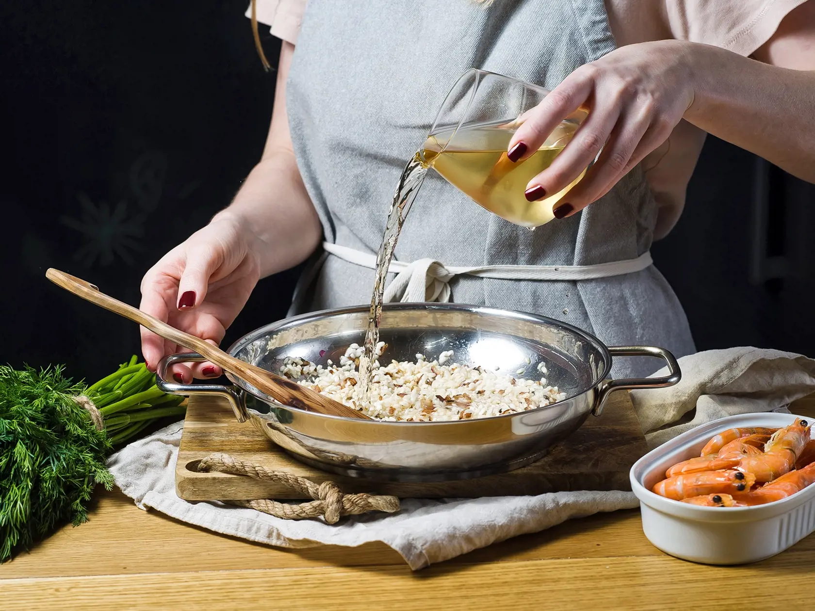 The chef pours white wine into an Italian risotto. Shrimp, white wine, rice, onions, thyme, garlic. Dark background, side view