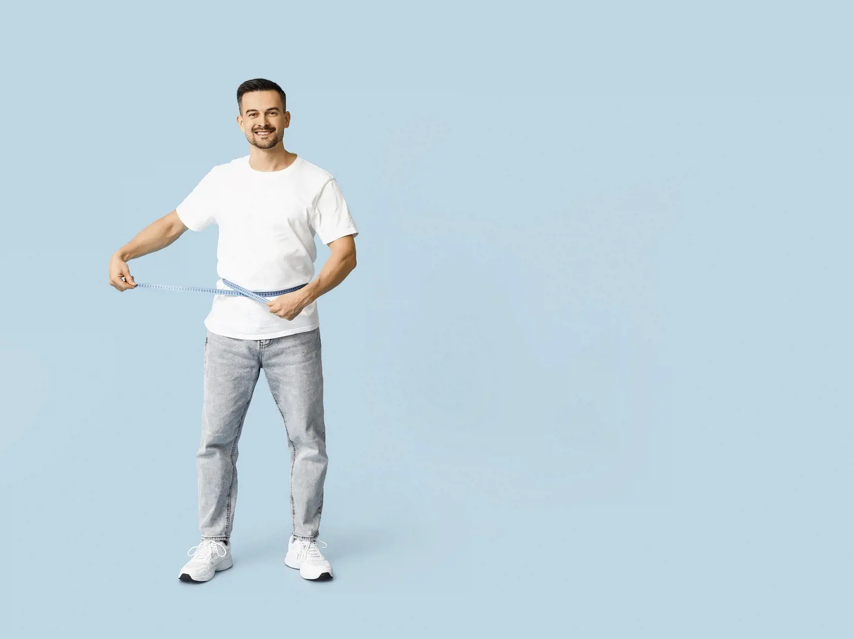 Handsome sporty happy man measuring his waist on blue background. Weight loss concept