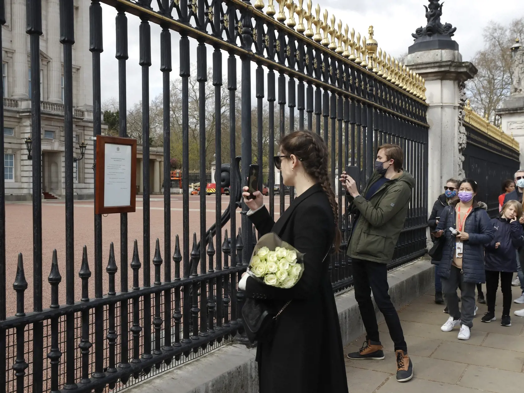KK4 Londýn - Žena si fotí oznámenie o úmrtí britského princa Philipa, ktoré visí na bráne Buckinghamského paláca 9. apríla 2021 v Londýne. Britský princ Philip, manžel kráľovnej Alžbety II., zomrel v piatok vo veku 99 rokov. Oznámil to Buckinghamský palác.  "Jeho kráľovská výsosť zomrela dnes ráno pokojne na Windsorskom zámku," dodal palác s tým, že ďalšie informácie zverejnia v náležitom čase. FOTO TASR/AP
A woman carries flowers as she takes a mobile phone photo of an announcement regarding th