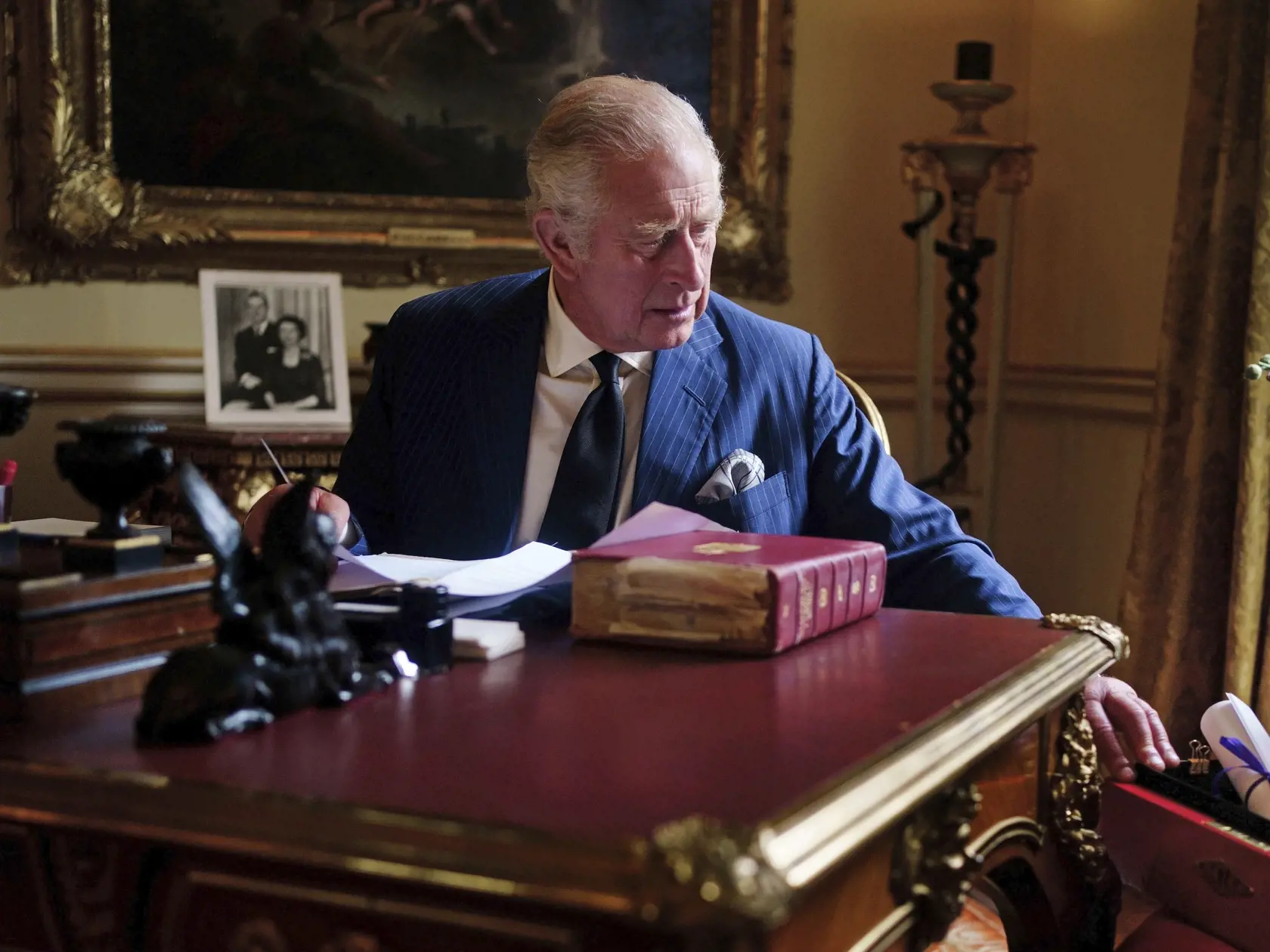 Na snímke nový britský kráľ Karol III. sedí za pracovným stolom a vyberá z oficiálnej červenej krabice korešpodenciu, ktorú mu pripravil osobný tajomník v miestnosti z 18. storočia v Buckinghamskom paláci 11. septembra 2022. ritská kráľovská rodina zverejnila prvú fotografiu nového kráľa pri plnení si štátnych povinností. Fotografia vznikla tri dni po smrti kráľovnej Alžbety II. Ako súčasť panovníckej kontinuity sú na fotografii za ním jeho zosnulí rodičia Alžbeta a Filip.