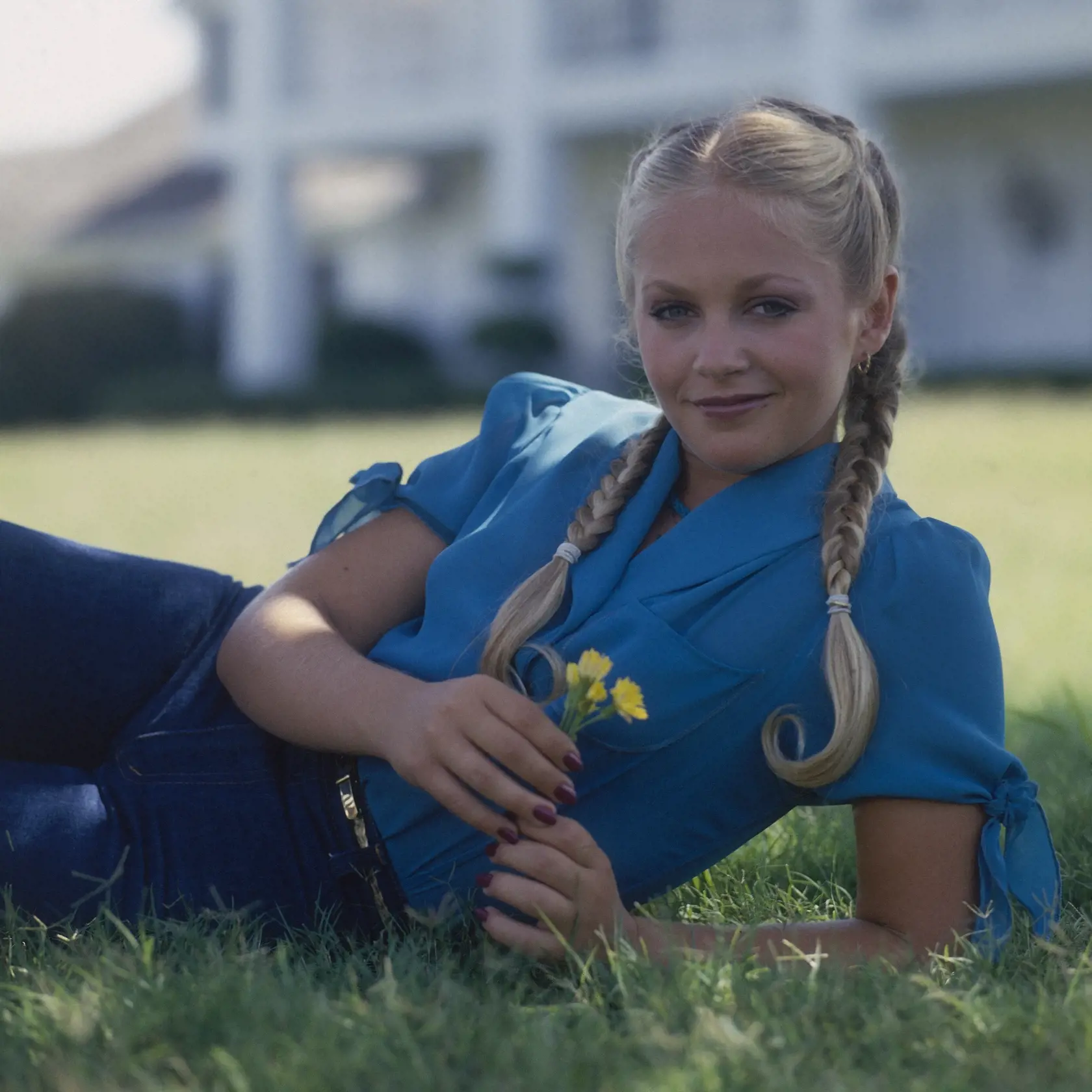 Promotional portrait of American actress Charlene Tilton, her hair in braids and flowers in her hands, as she lies on the grass in front of the Southfork Ranch, the television home of the Ewing family from the series 'Dallas,' Dallas, Texas, 1979. Tilton portrayed Lucy Ewing on the long-running series.(Photo by CBS Photo Archive/Getty Images)
