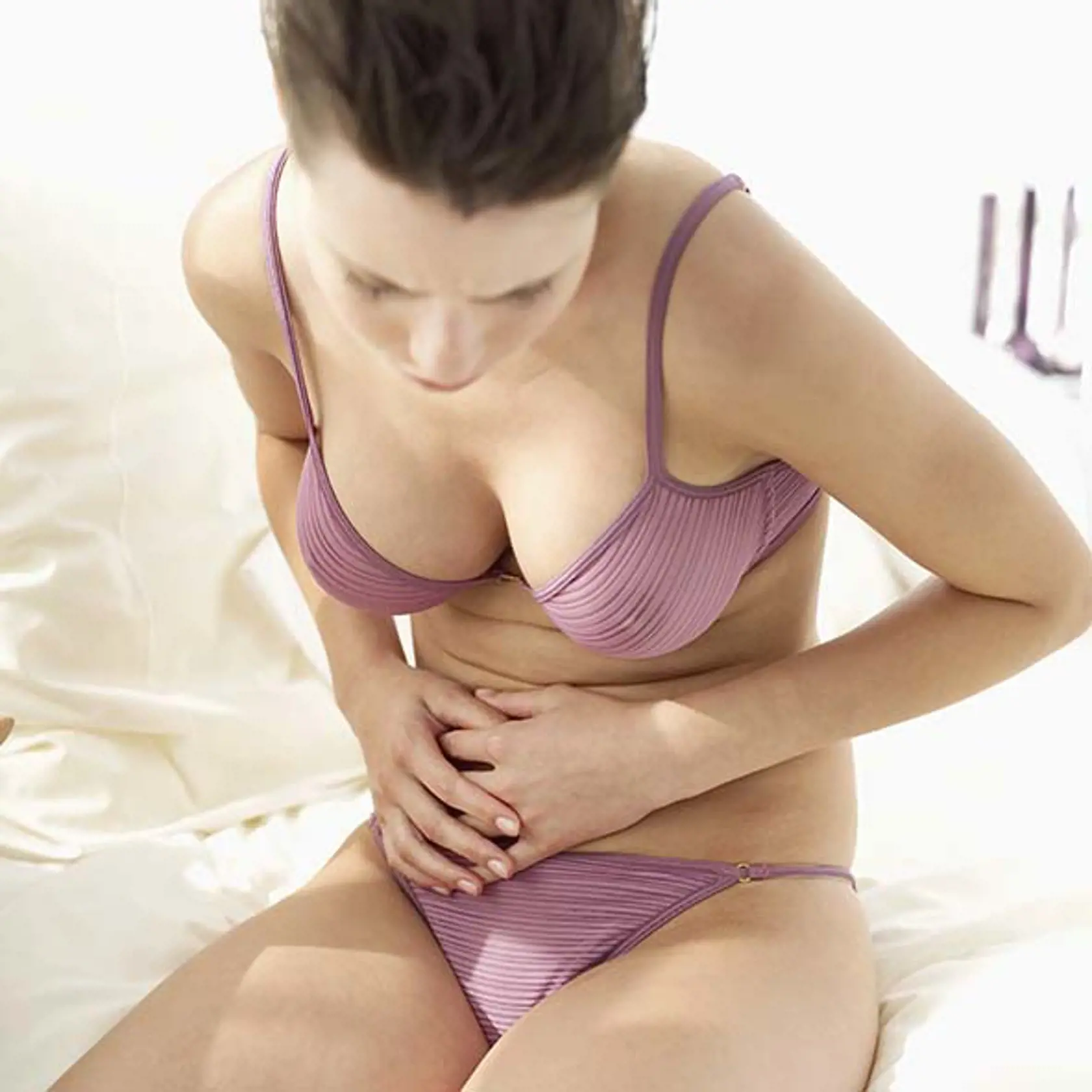 Young woman sitting on the edge of a bed wearing underwear and holding her stomach