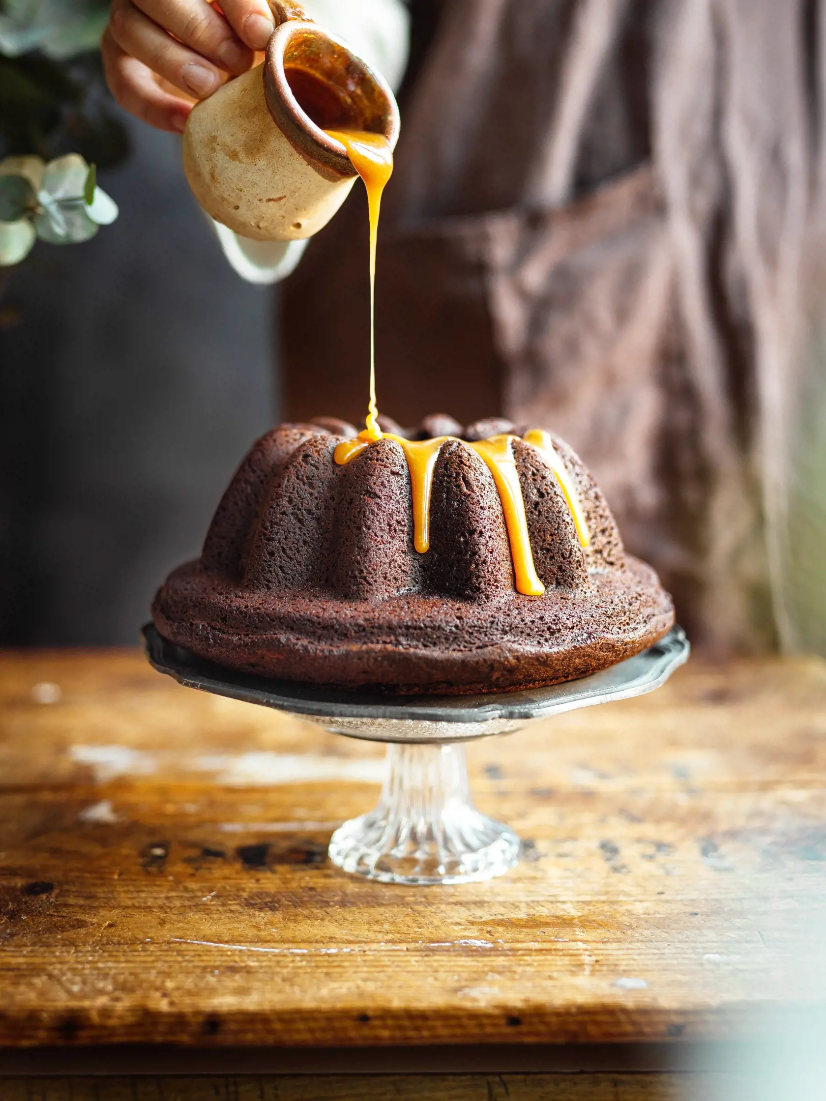 Person,In,The,Background,Pouring,Caramel,Over,Chocolate,Bundt,Cake.
