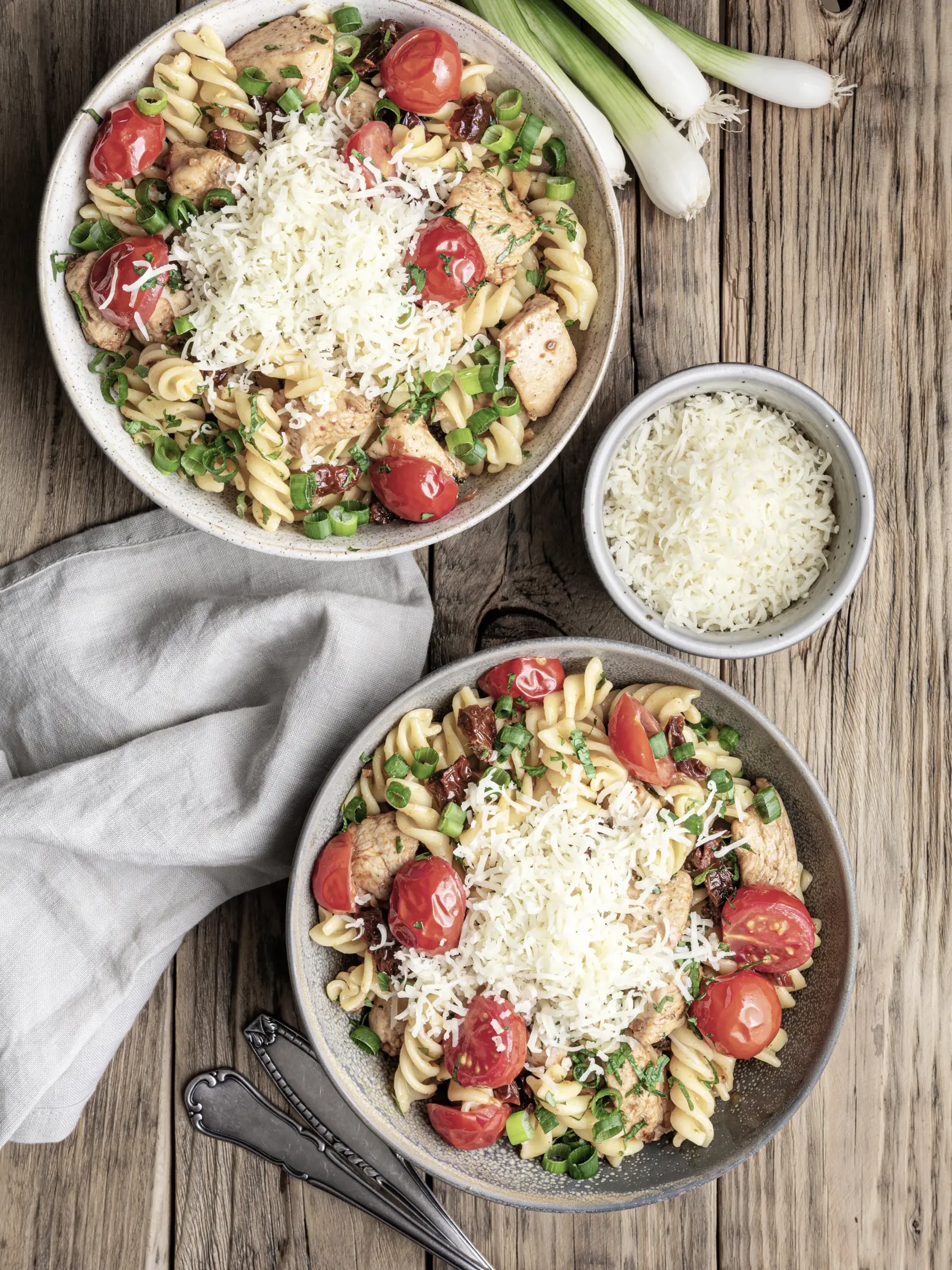 Chicken pasta salad with roasted and sun-dried tomatoes, topped with green onion slices and grated cheese on wooden background