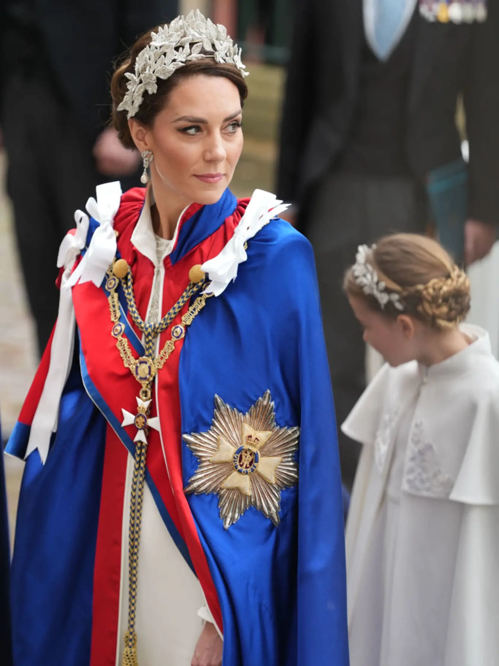 Princezná Kate mala na sebe tradičný ceremoniálny plášť. Pod ním slonovinové šaty od Alexandra McQueena a 3D čelenku, rovnako tak aj jej dcéra, princezná Charlotte.