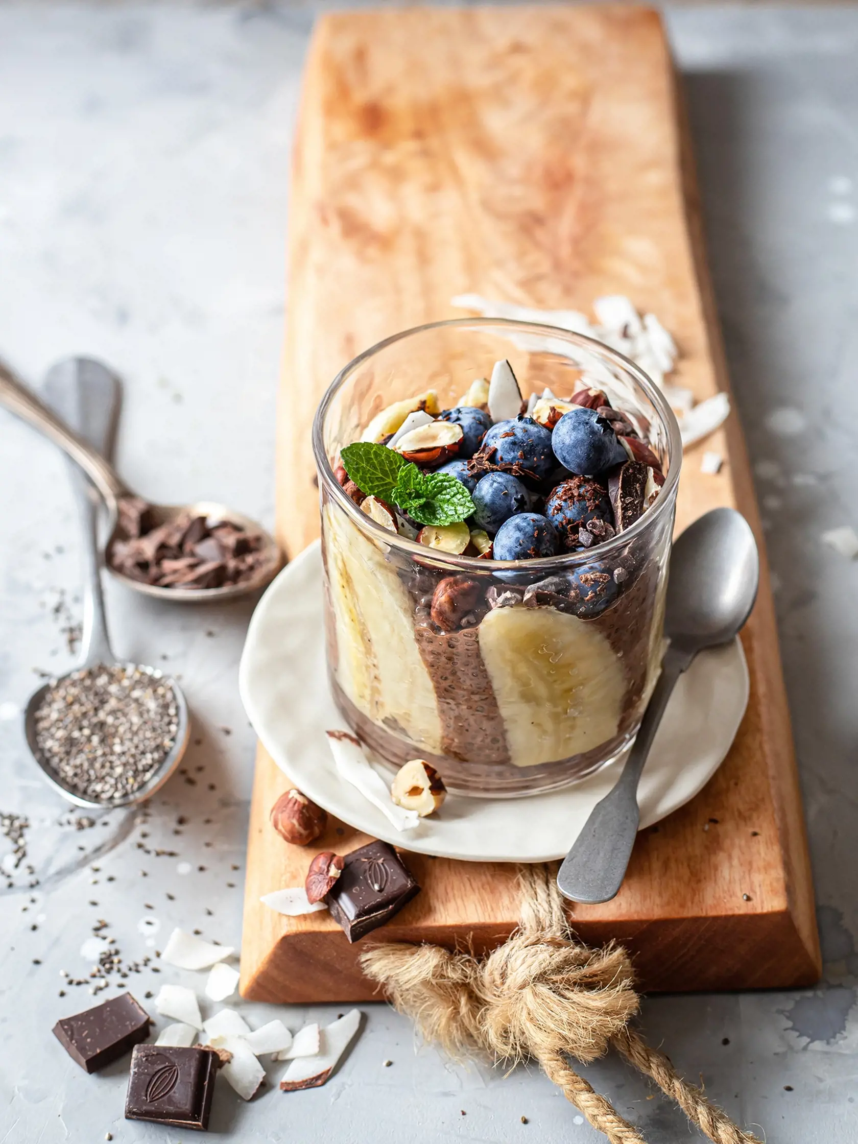 Raw Chocolate overnight Chia Seeds Pudding made with coconut milk, served with banana slices, cream, hazelnuts, fresh blueberries, peppermint leaves and chocolate chips.  Close up.