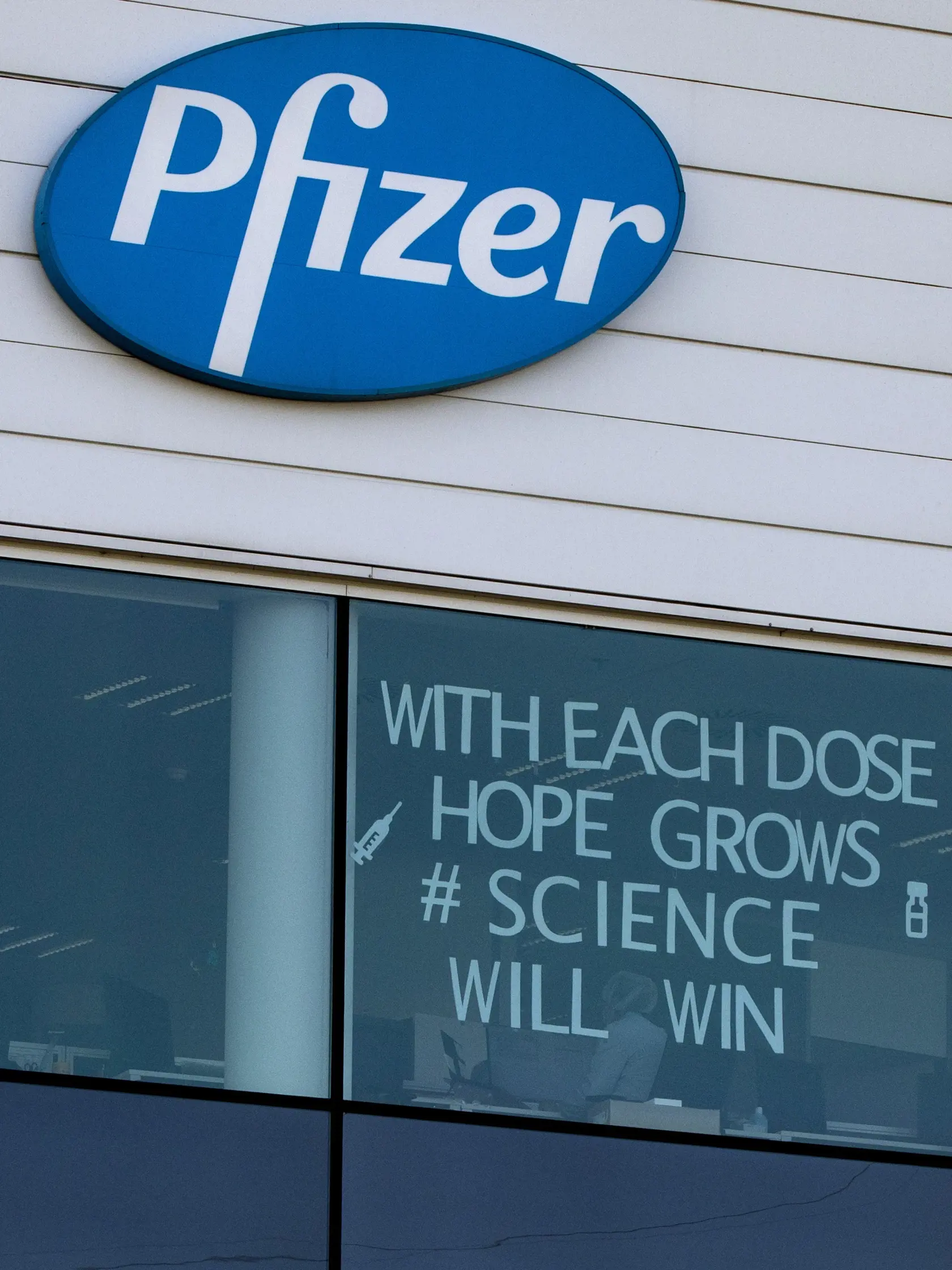 KK19 Puurs - Na snímke logo Pfizeru vo výrobnom centre spoločnosti v belgickom meste Puurs v utorok 23. februára 2021. Farmaceutické spoločnosti Pfizer a BioNTech vo štvrtok 25. februára 2021 oznámili, že skúmajú možnosť pridania tretej dávky vakcíny proti koronavírusu, ktorá sa momentálne podáva v dvoch dávkach v rozstupe niekoľkých týždňov. Takisto testujú novú verziu svojej vakcíny, ktorá sa má zamerať na juhoafrický variant vírusu. FOTO TASR/AP
An employee works behind a window with a messag
