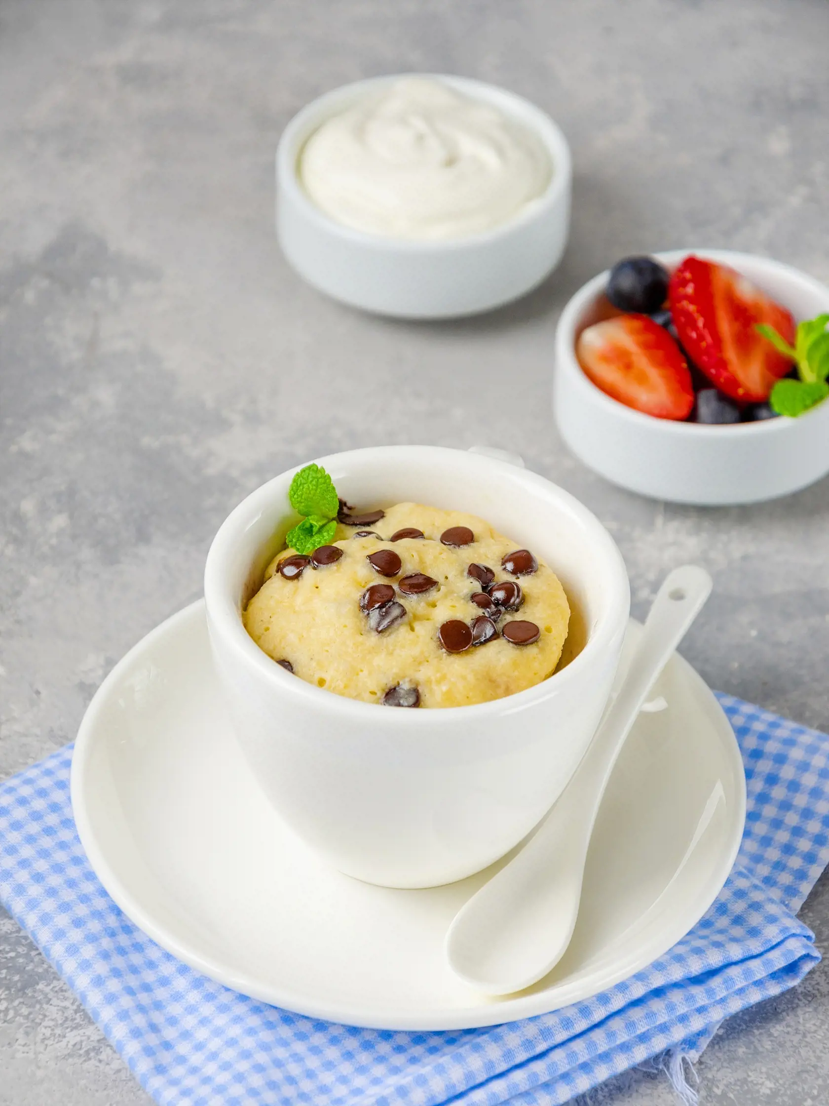 Mug cake with chocolate chips, fresh berries and cream on a gray concrete background. Copy space