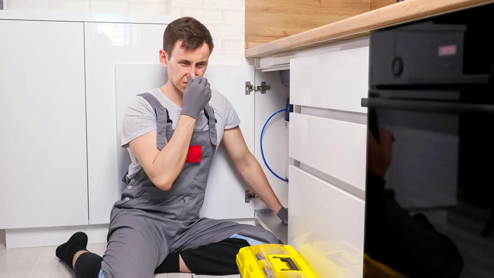 Disgusted young plumber in grey uniform and gloves sits on floor and holds nose from smell repairing waste trap under sink in light kitchen