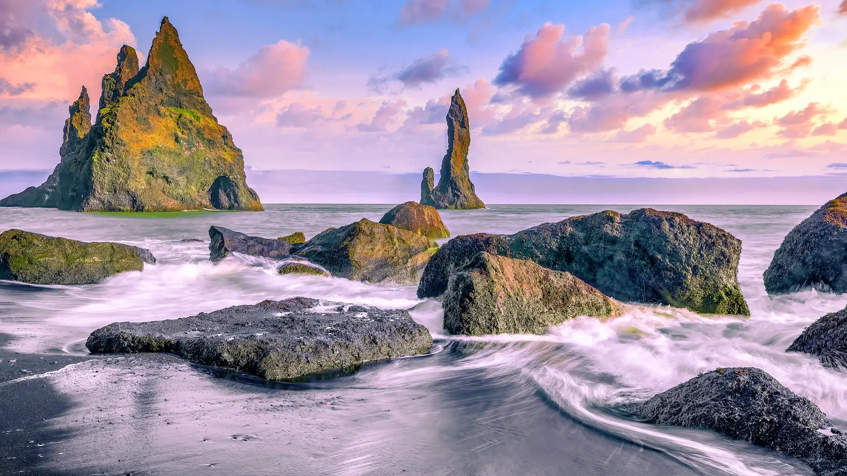 Čadičové skaly – Reynisfjara. Mohutné tmavé stĺpy čadiča sa týčia z čiernej piesočnatej pláže, kde rozbúrené vlny Atlantiku narážajú na monumentálne skalné útvary, vytvárajúc kontrast medzi silou prírody a krásou pobrežia.