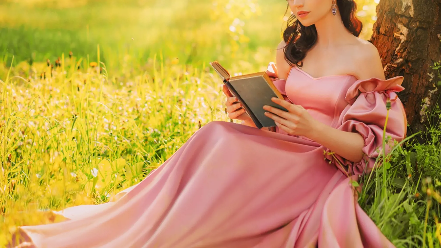 Fantasy woman sits under tree holding romantic book in hands reading novel smiling face, lips close-up. Pink long dress. Fairy girl in summer nature green grass magic sun light. art photo real people