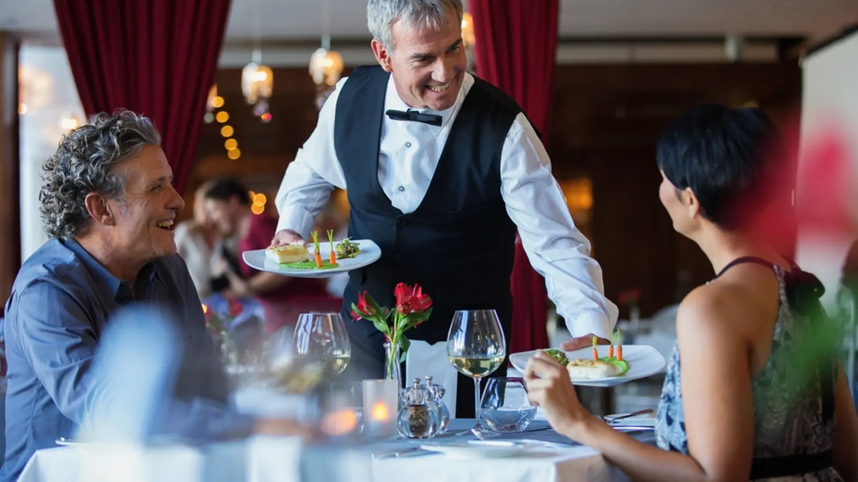 Smiling waiter serving fancy dishes to mature couple sitting at table in restaurant