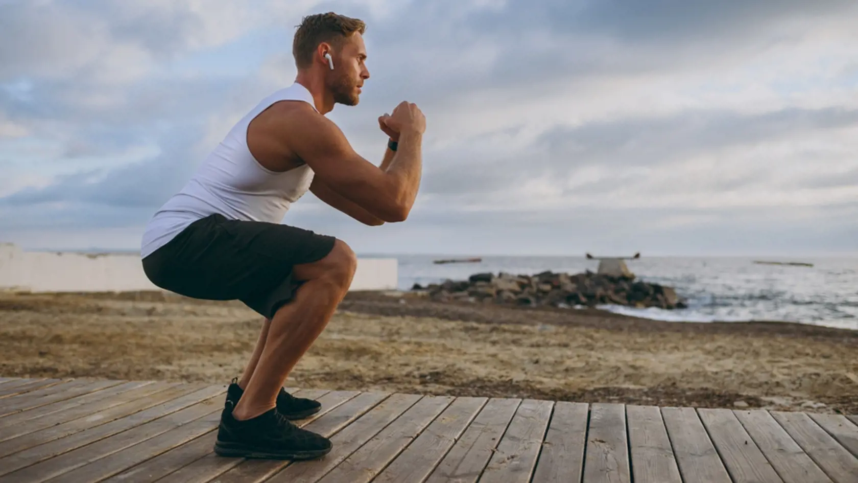 Full body young strong sporty athletic toned fit sportsman man wears sports clothes earphones do squats warm up train at sunrise sun dawn over sea beach outdoor on pier seaside in summer day morning