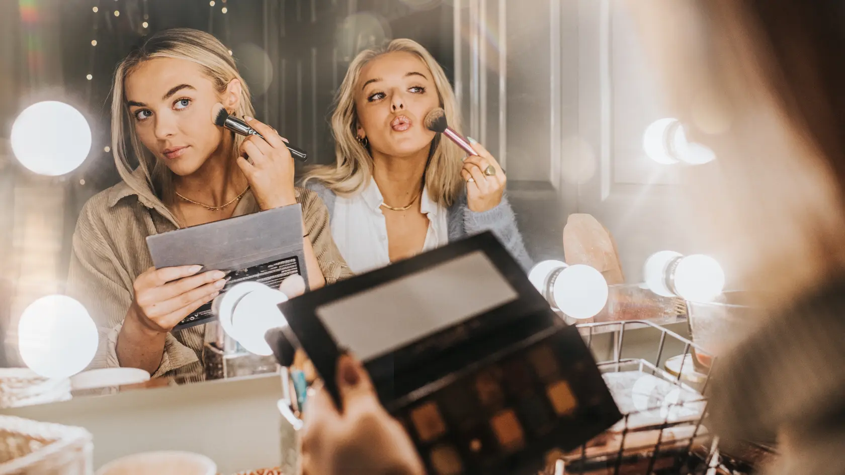 Two young woman in their twenties sit at a vanity table by a mirror surrounded by bulbs. They concentrate as they apply make-up to their faces with brushes. They are comfortable with each other. The scene is carefree and youthful as the lights create flare and reflections.